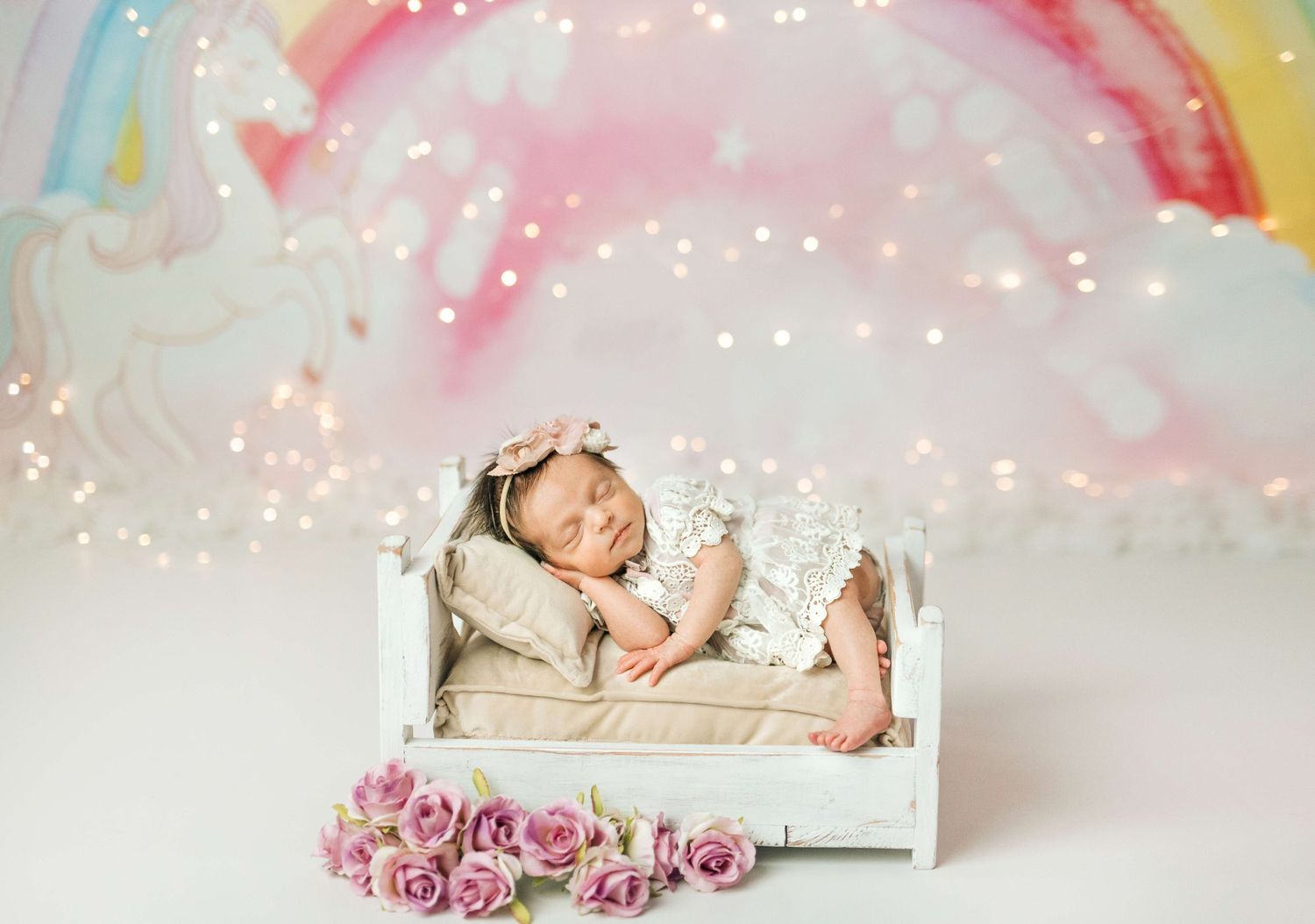 A sleeping newborn in a white lace outfit rests on a tiny bed with pink roses and a rainbow unicorn backdrop.