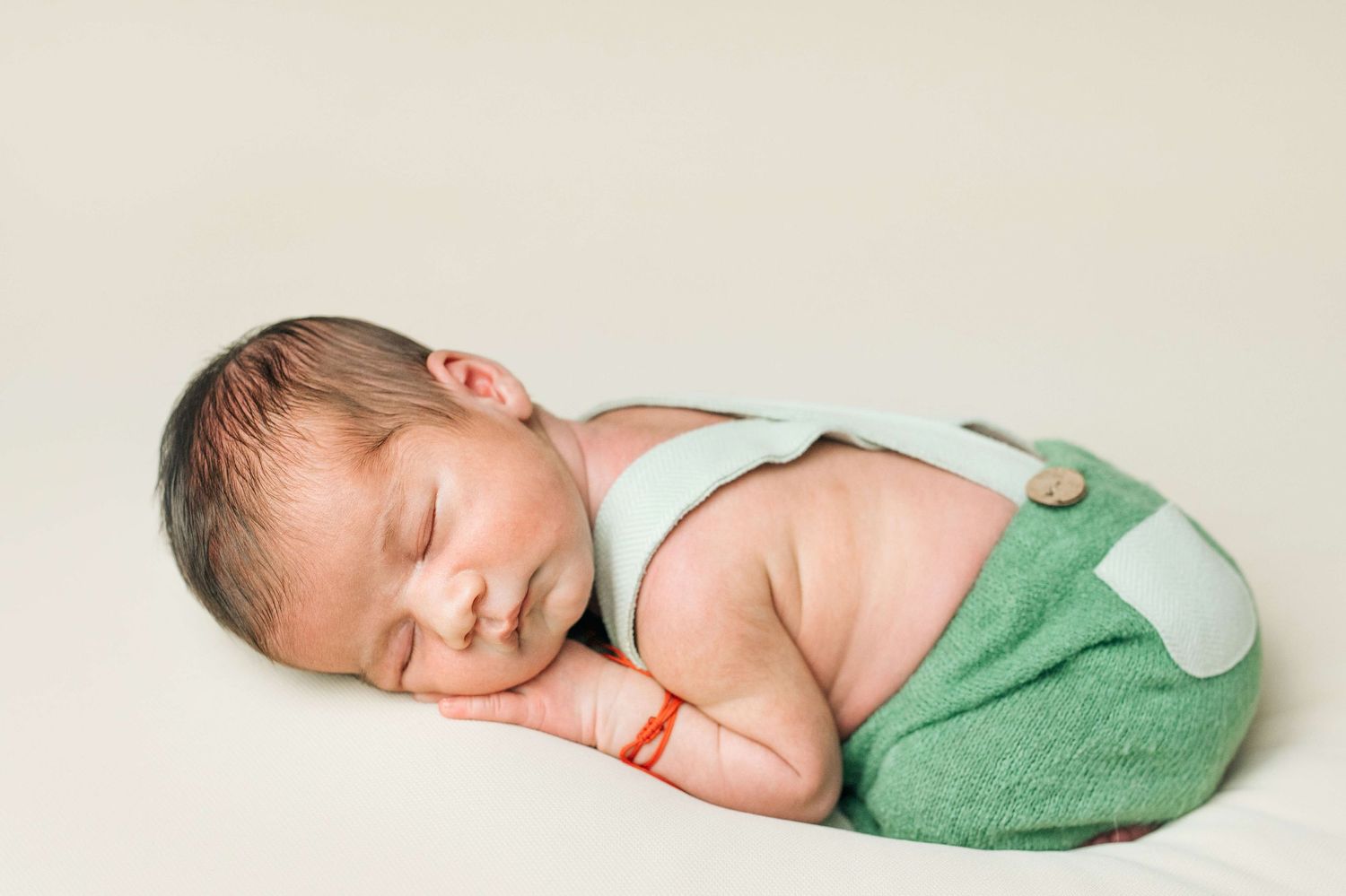 Sleeping newborn baby in green overalls and mint suspenders posed on a cream background.