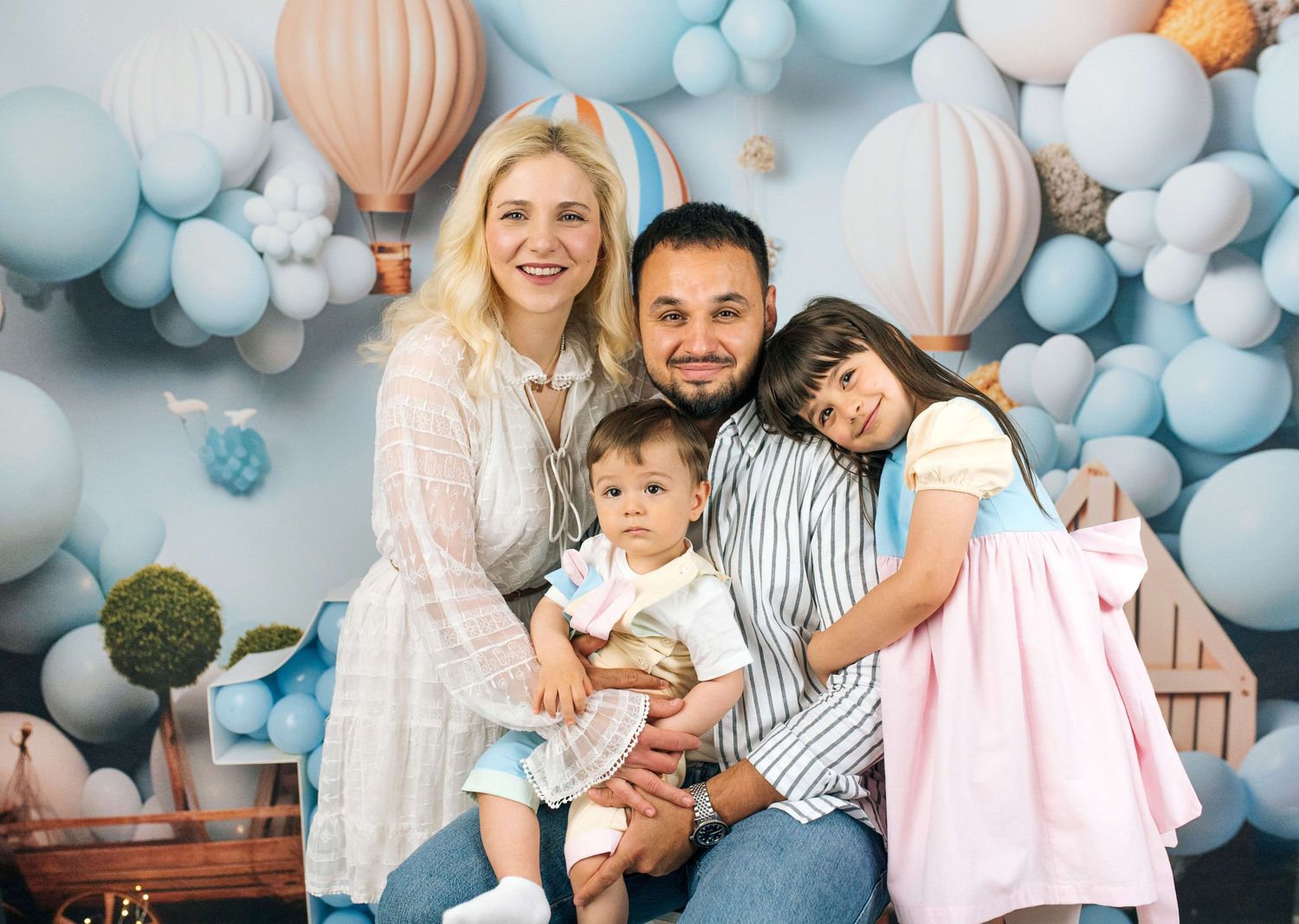 Family of four posing joyfully in front of a blue balloon wall with hot air balloon decorations.