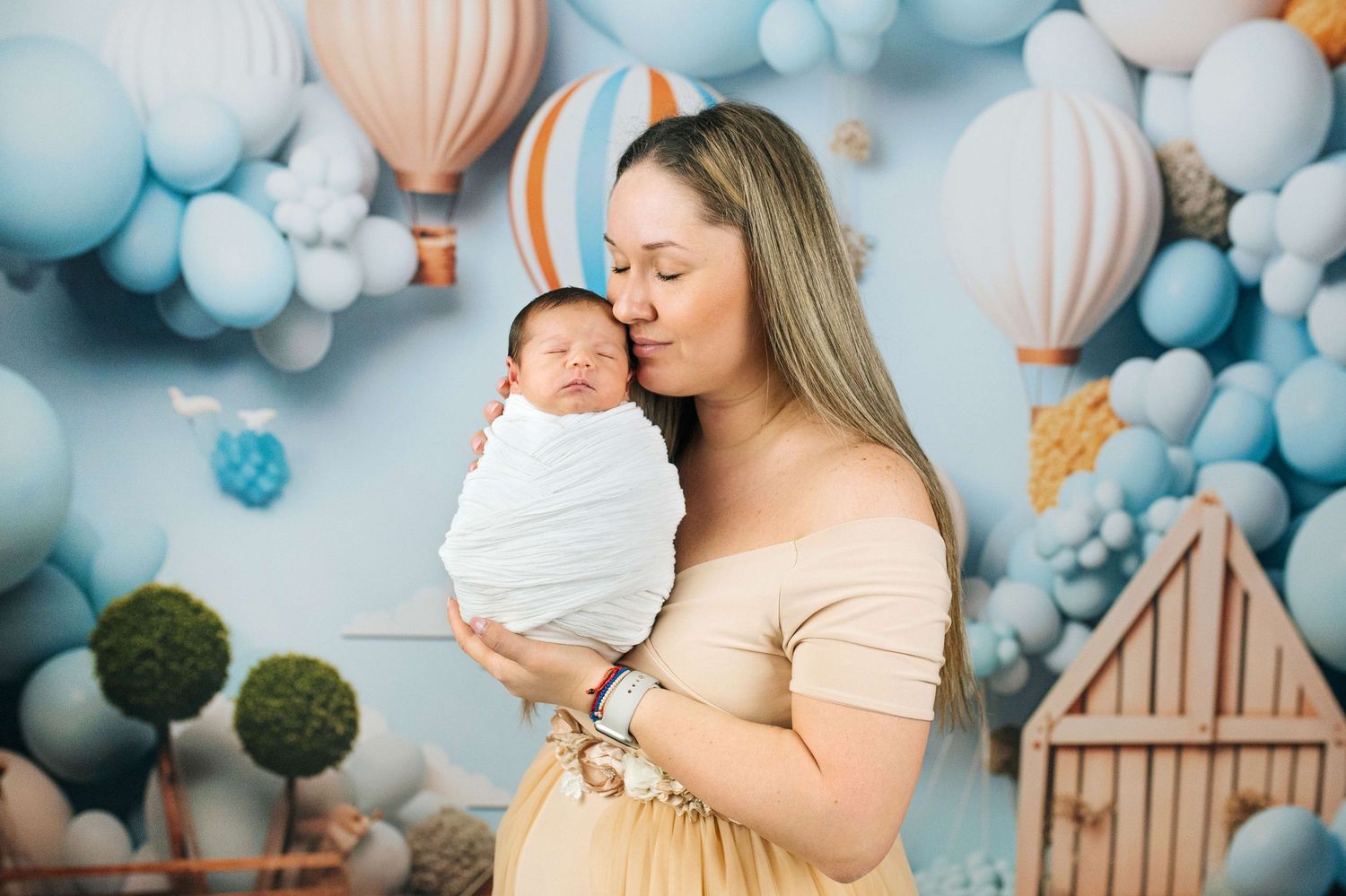A mother tenderly kisses her swaddled newborn baby against a dreamy blue balloon and hot air balloon backdrop.
