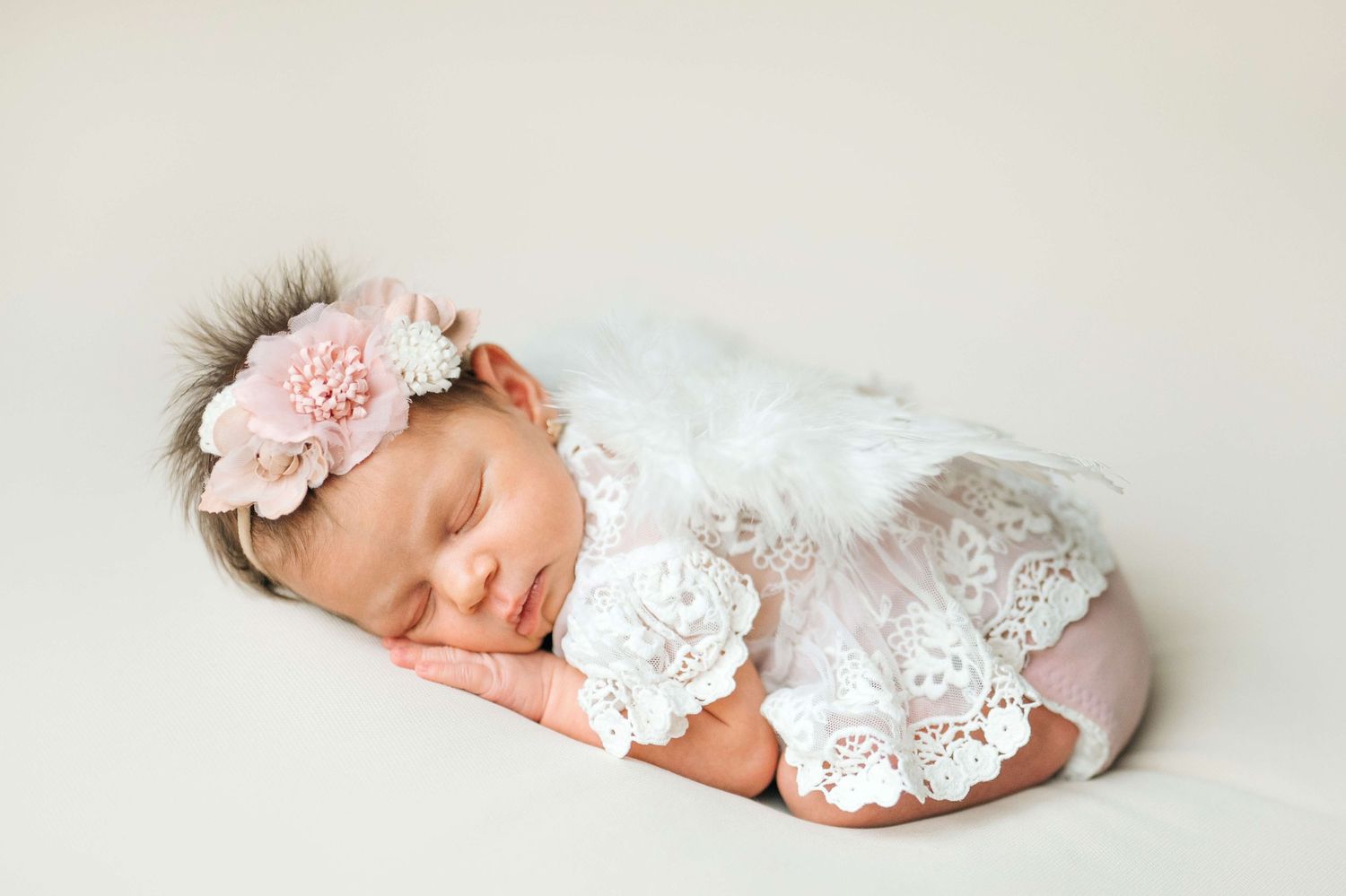 Sleeping newborn baby girl in white lace outfit and pink floral headband on cream background.