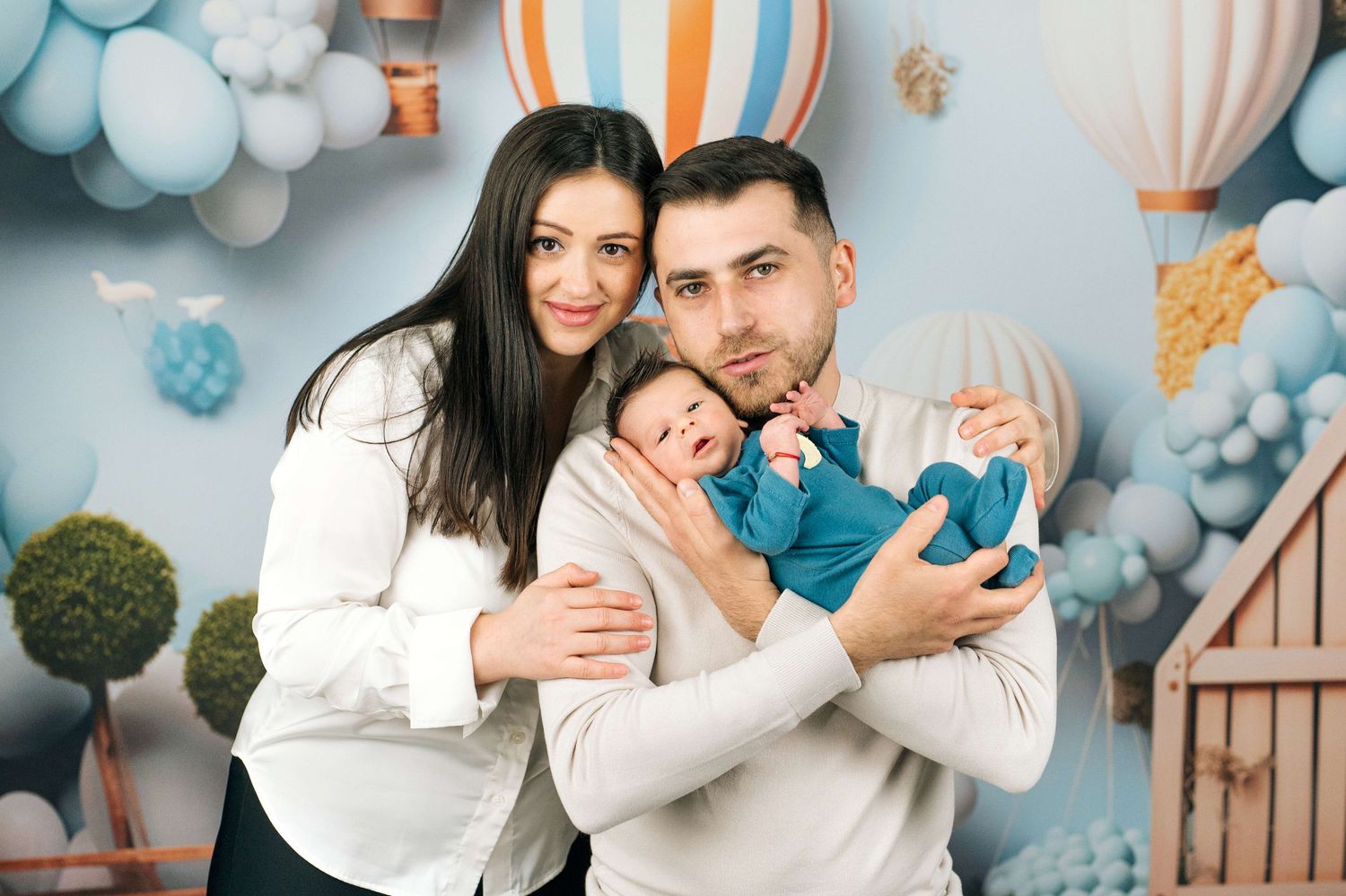 Happy parents holding their newborn baby in a blue onesie in front of a balloon-themed backdrop.