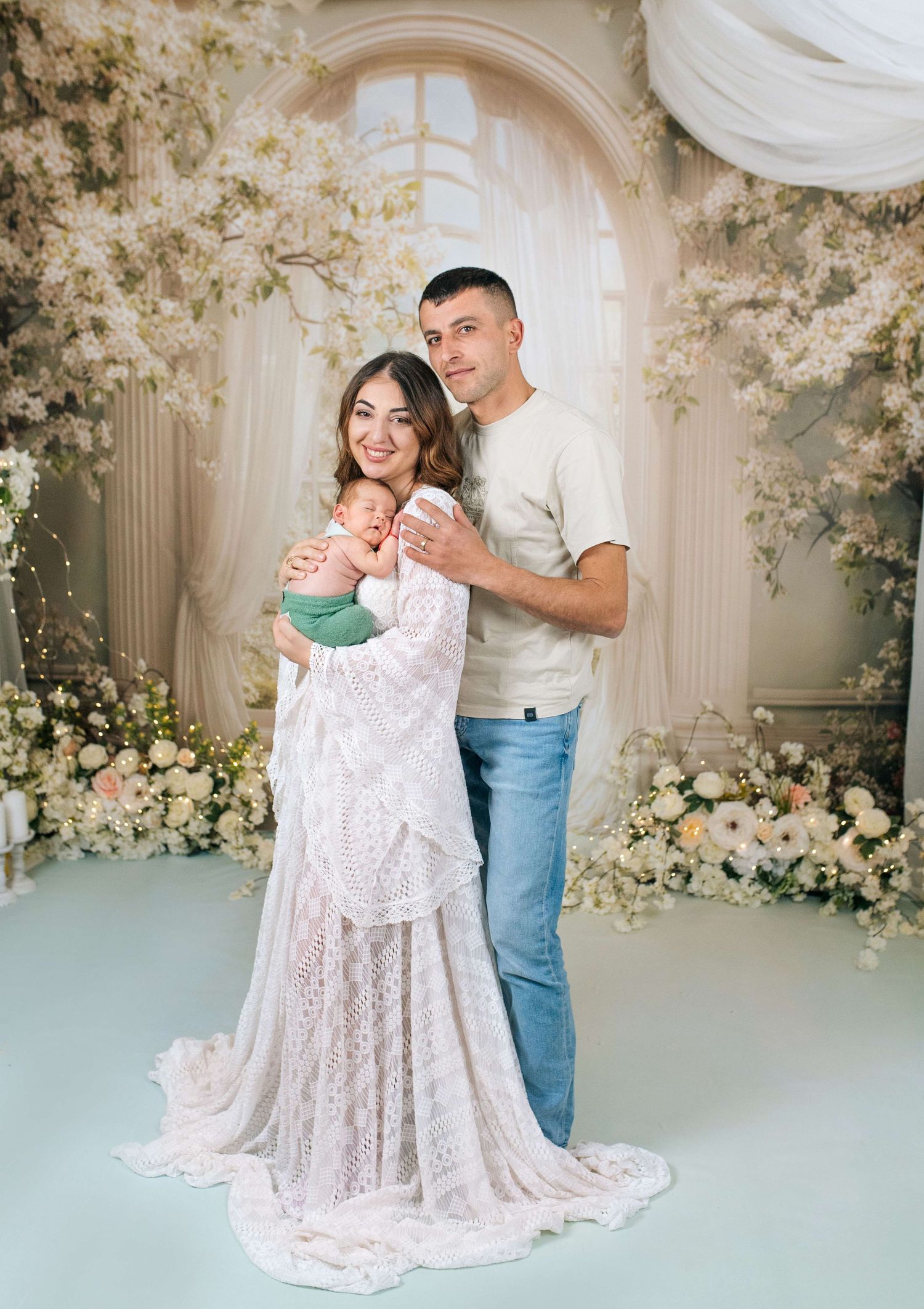 Young family posing with newborn baby, mother in white lace gown, father in casual attire, floral studio backdrop.