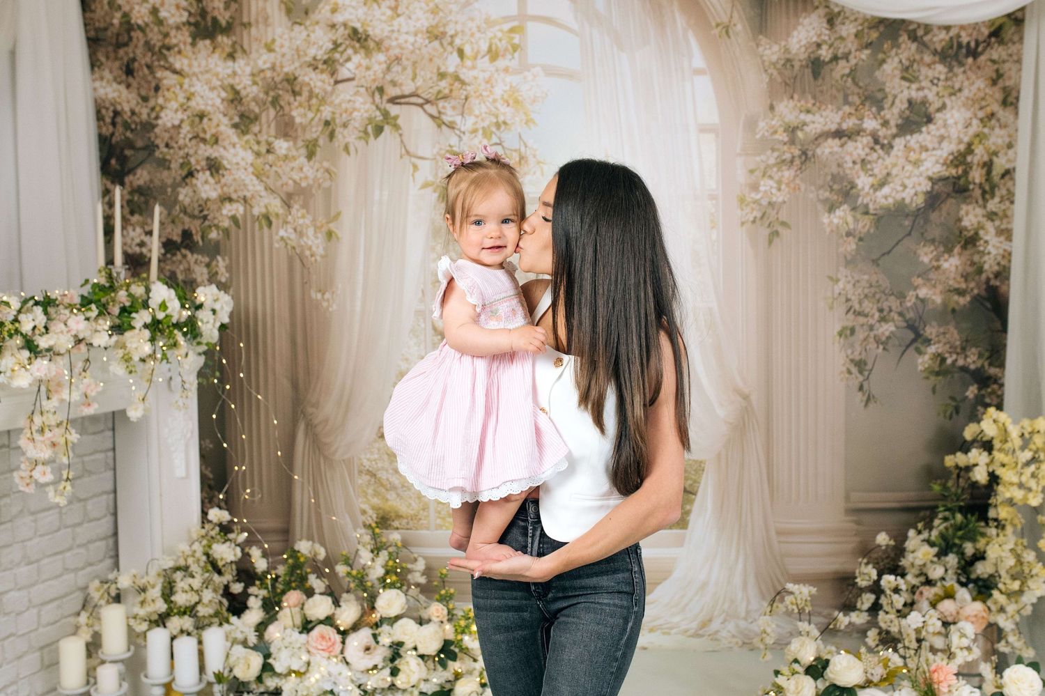 A mother in jeans kisses her toddler daughter in a pink dress amid elegant floral decorations.