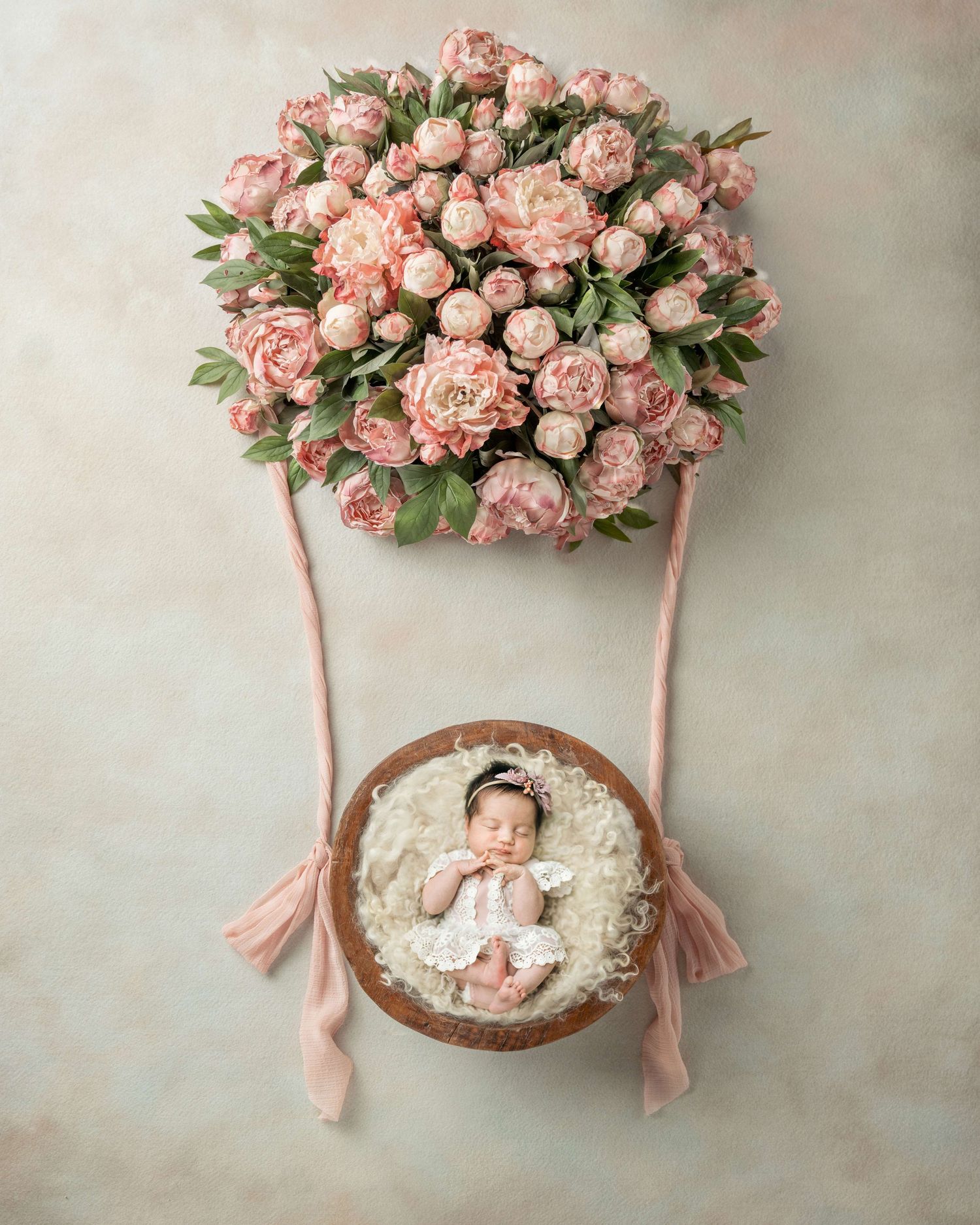 Newborn baby sleeping in a wooden bowl beneath a pink peony hot air balloon arrangement on a soft background.