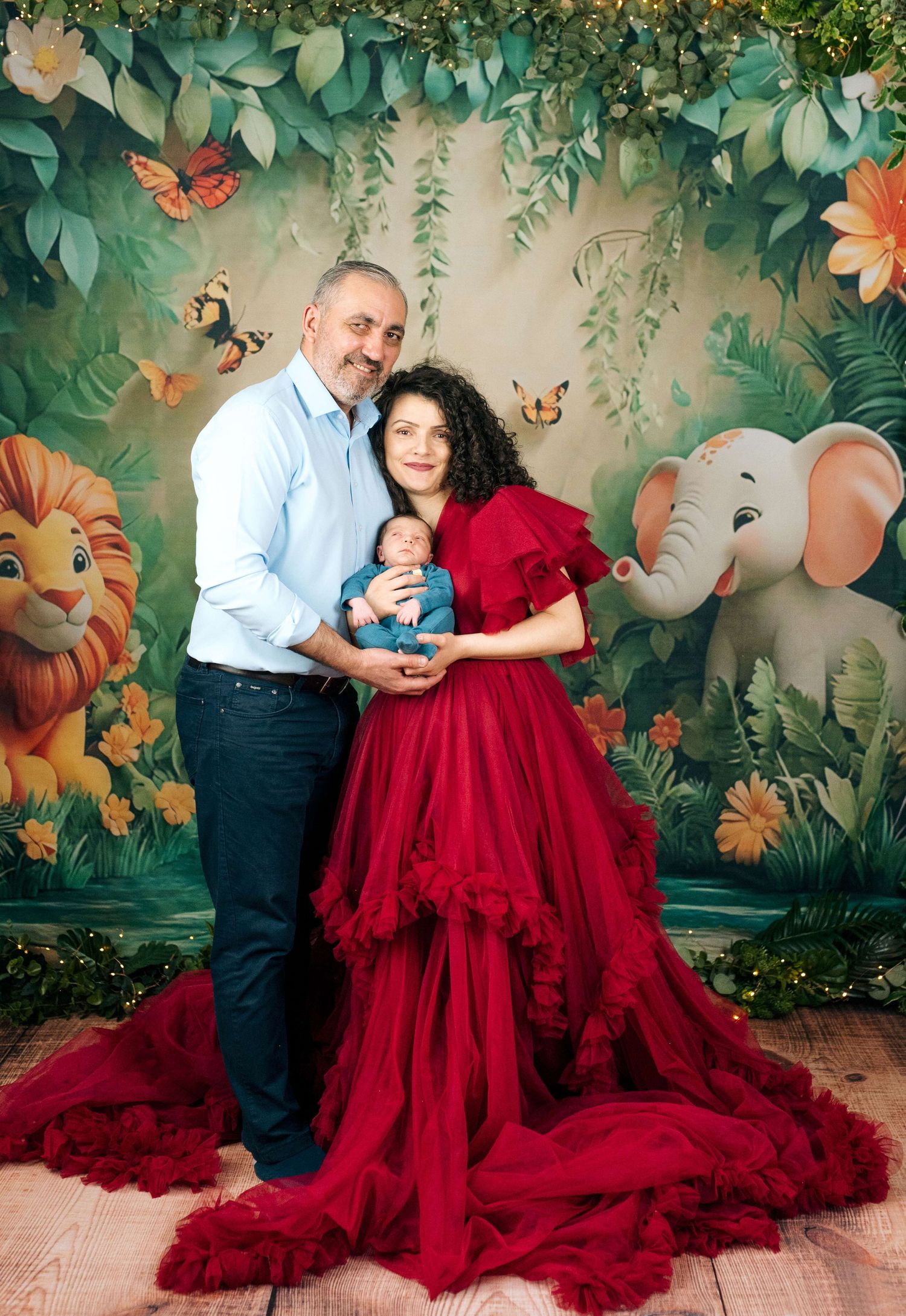 Family portrait with newborn baby, mother in red gown, father in blue shirt, jungle animal backdrop.