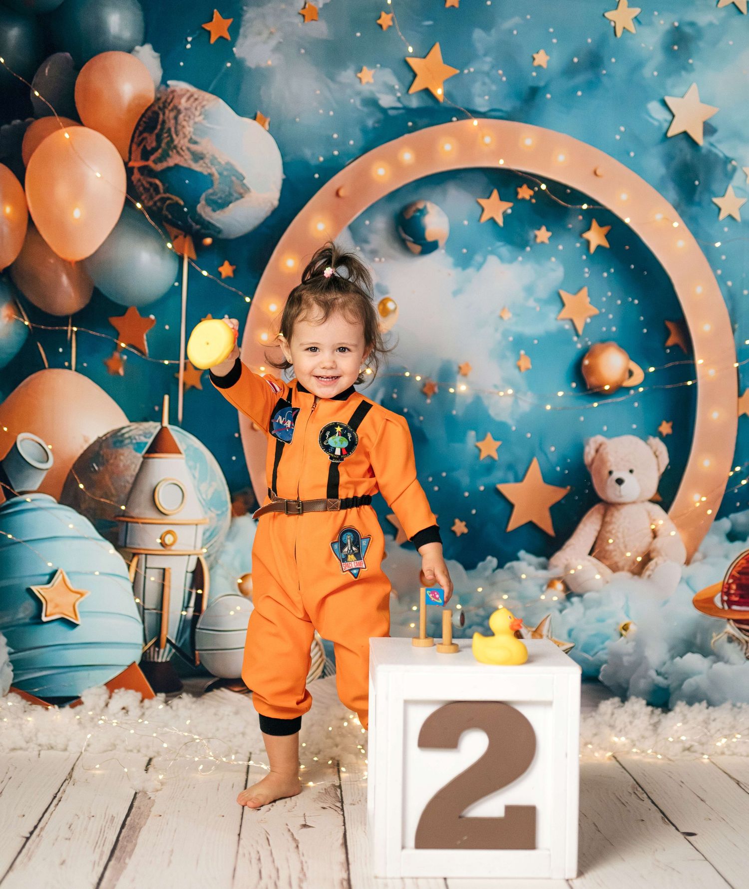 Toddler in orange astronaut jumpsuit poses by a number 2 block with space-themed balloons, stars, and rocket backdrop.