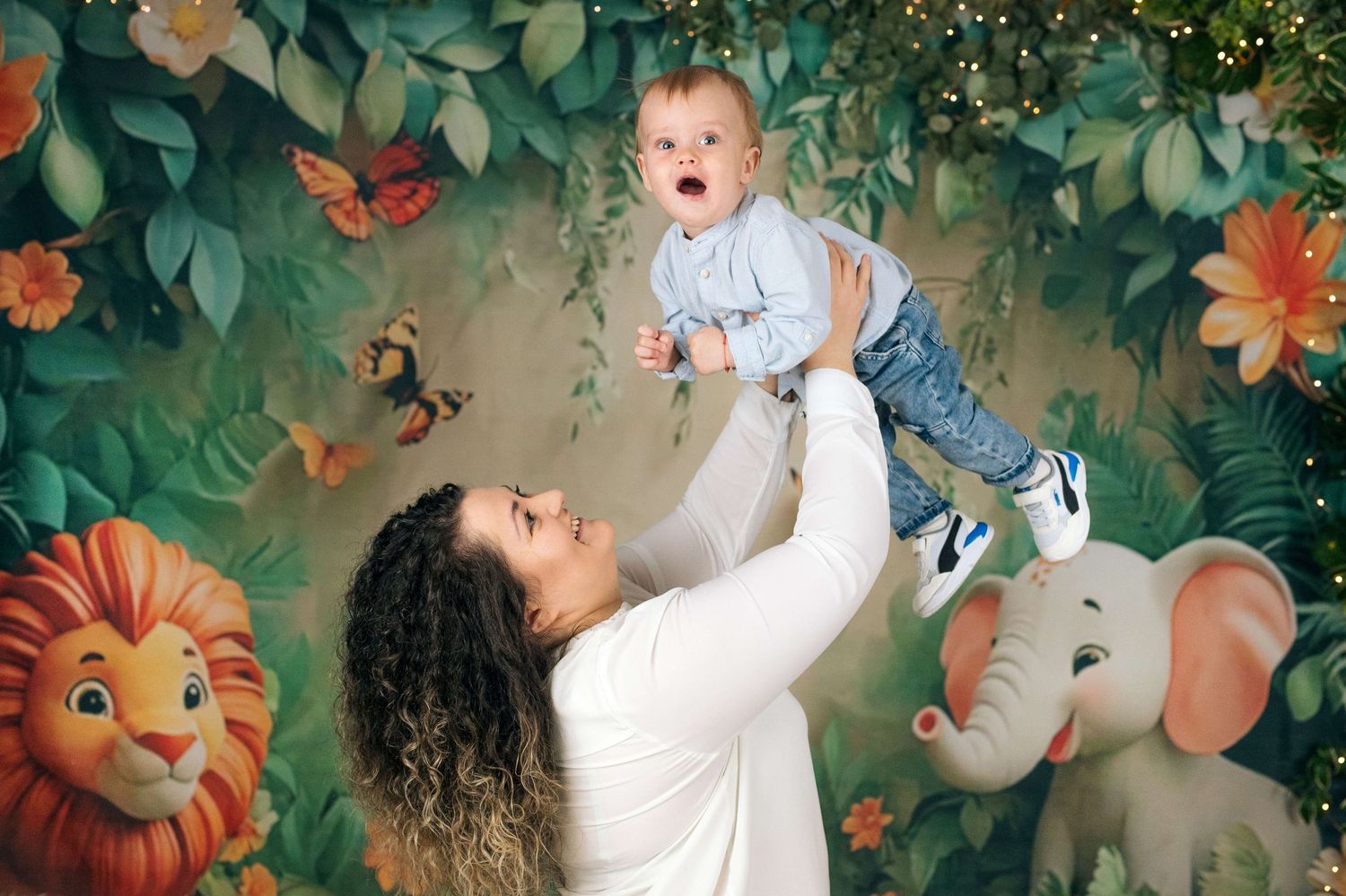 Mother lifting laughing baby boy high in air before vibrant jungle themed photo backdrop.