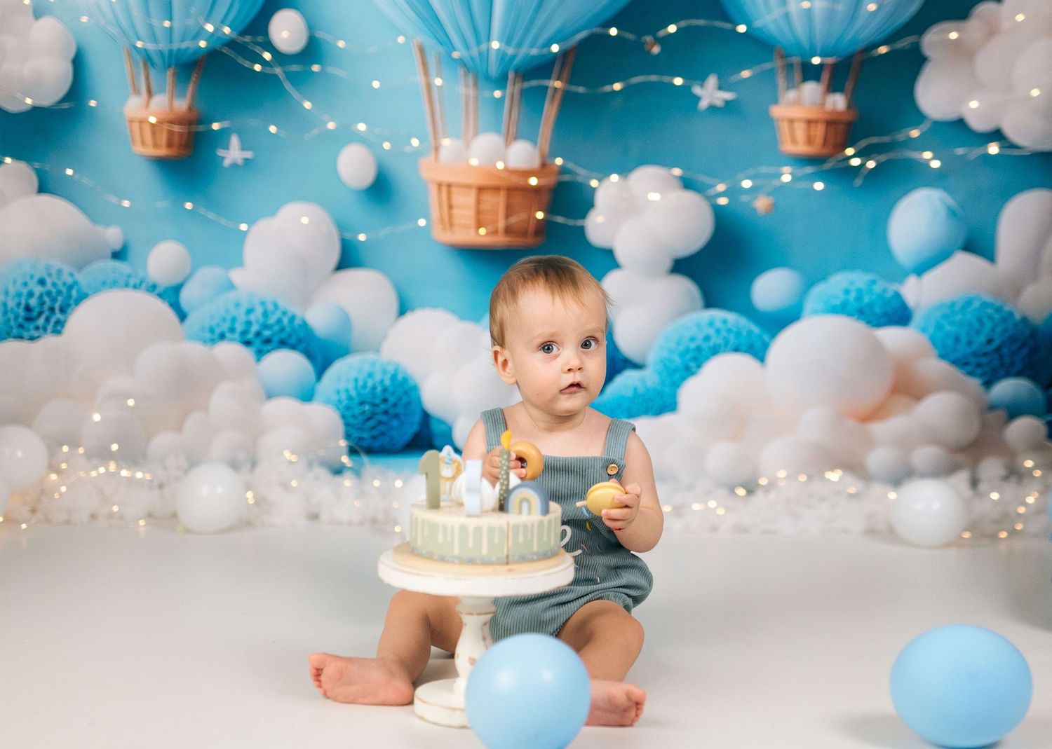 Baby boy in denim romper sits before a white birthday cake, surrounded by blue balloons and hot air balloon backdrop.