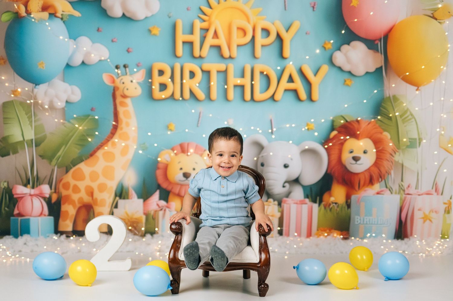 Smiling toddler sits in chair at safari-themed 2nd birthday party with giraffe, elephant, lion, balloons, and Happy Birthday banner.