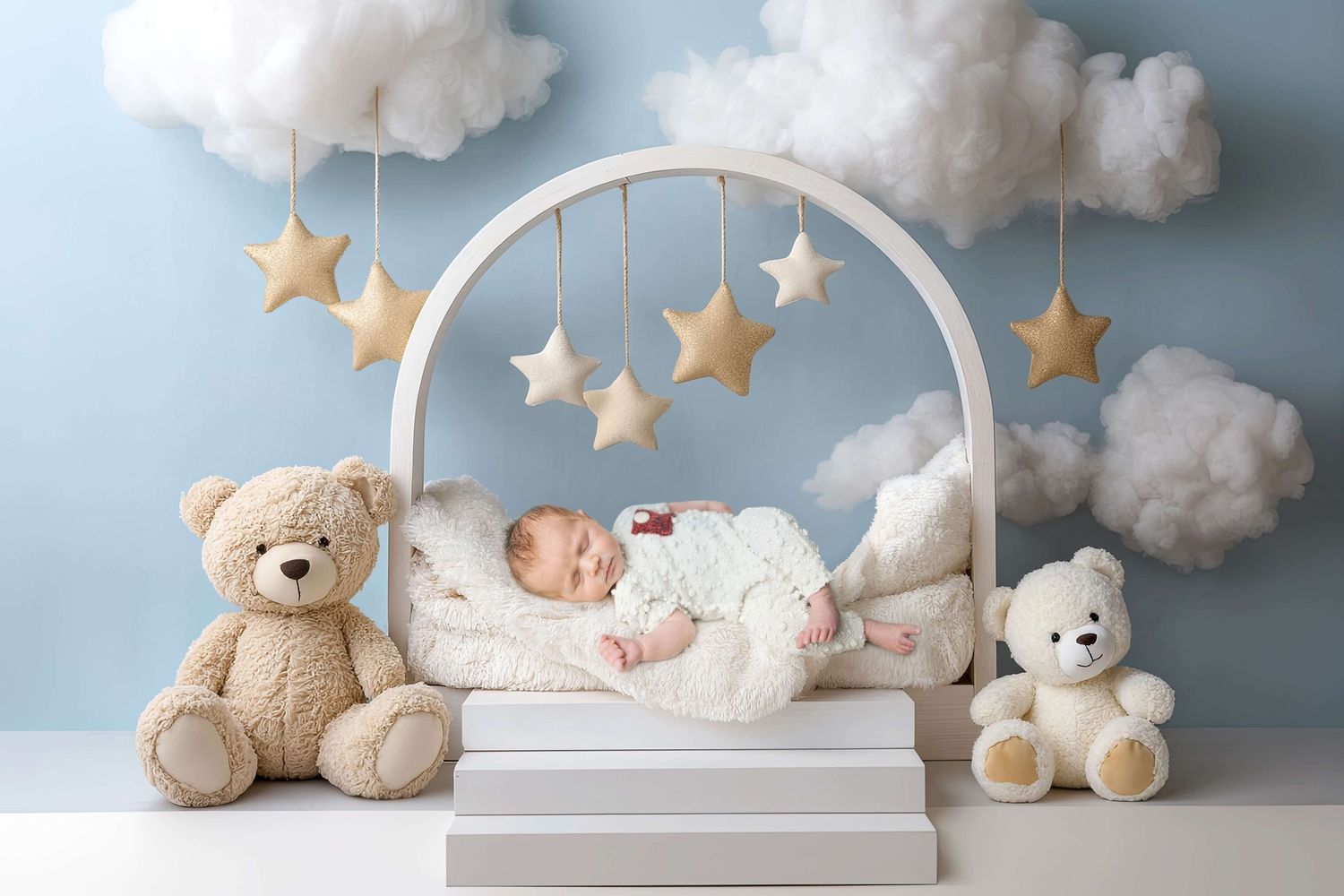 Newborn baby sleeping in white arch crib with teddy bears, star mobiles, and fluffy clouds on blue nursery backdrop.