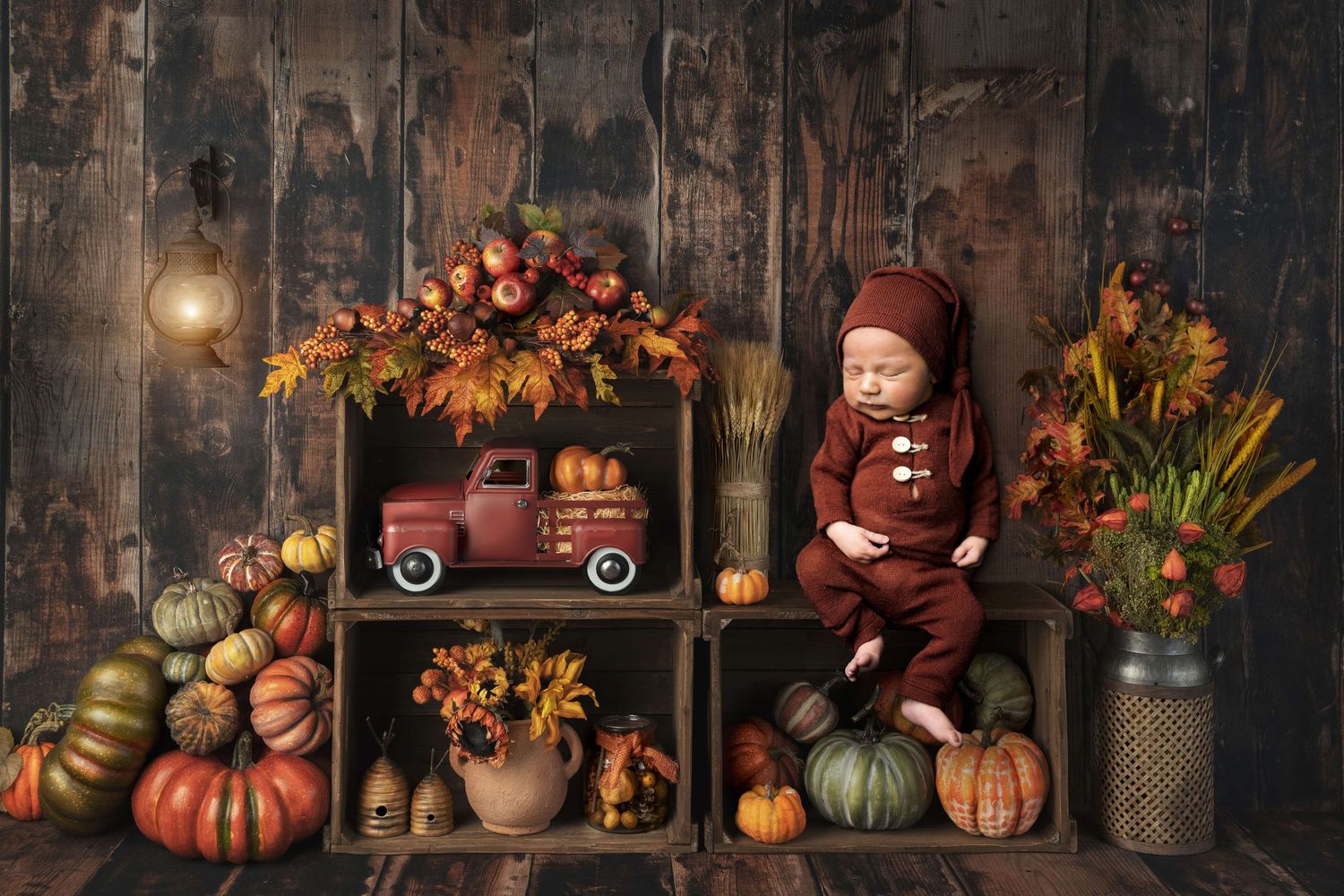 Baby in burgundy outfit sits among fall pumpkins, wooden crates, autumn foliage, and a red toy truck in a rustic setting.