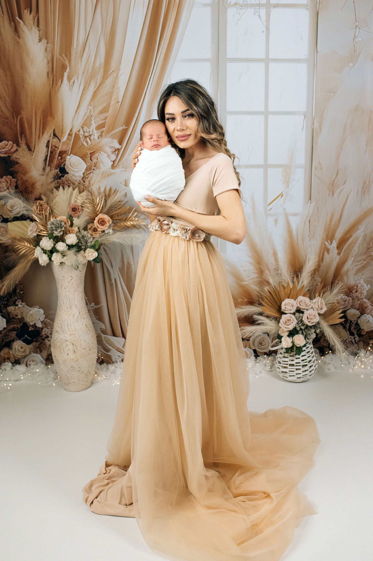 Elegant woman in gold tulle gown holding swaddled newborn baby, surrounded by boho floral decor.