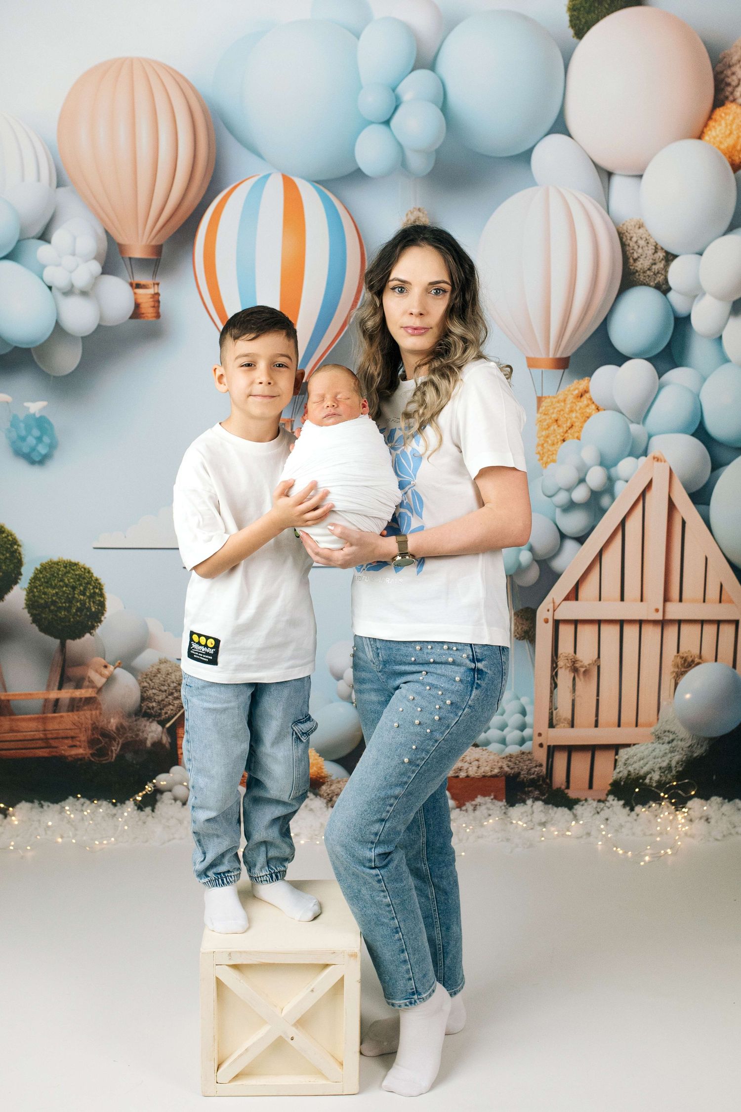 A mother in white tee and jeans holds newborn baby beside older son, posing before colorful balloon backdrop.
