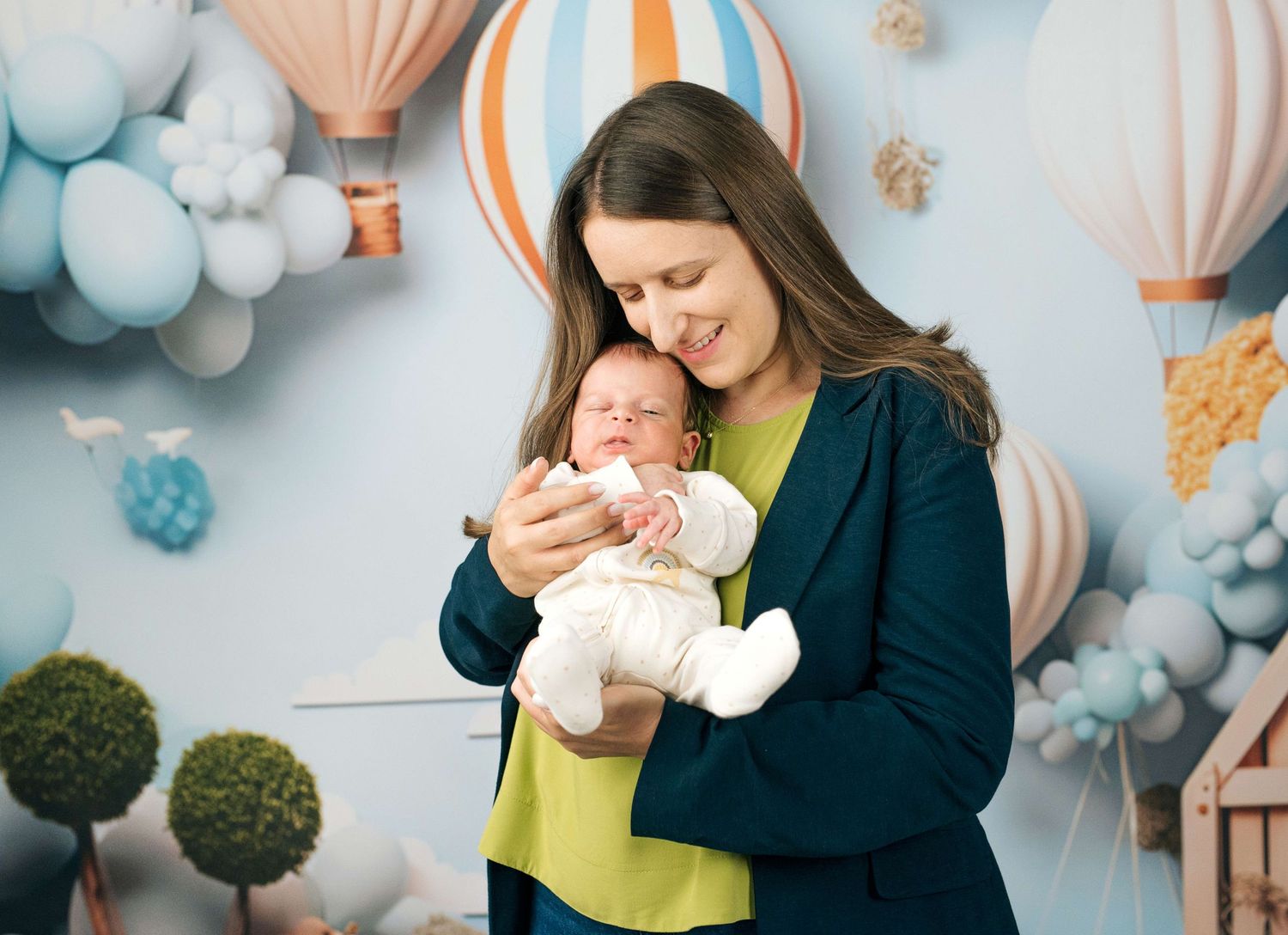 A smiling mother in a teal blazer cradles her newborn baby in a whimsical hot air balloon themed studio.
