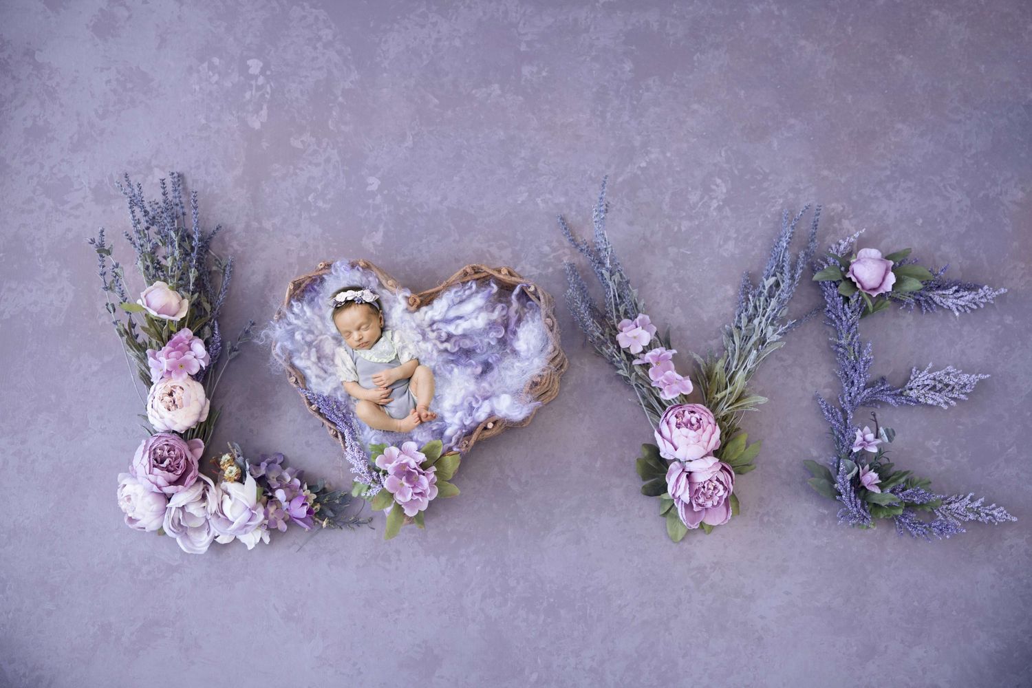 Newborn baby sleeping in heart-shaped nest surrounded by floral LOVE letters in purple and pink tones.