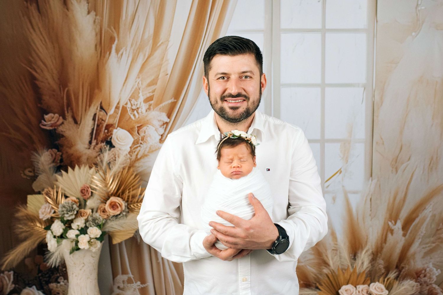 Smiling man in white shirt holding swaddled newborn baby with floral headband in boho-styled photo studio.