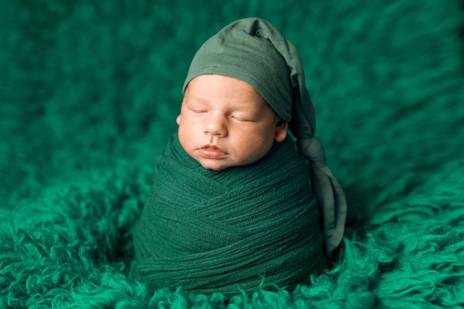 Newborn baby wrapped in green fabric with matching hat, sleeping peacefully on a fluffy green blanket.