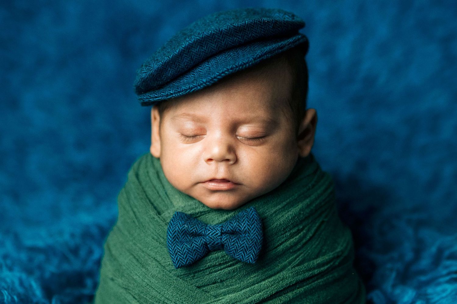 Sleeping newborn baby wrapped in green swaddle wearing a blue flat cap and bow tie on a blue background.