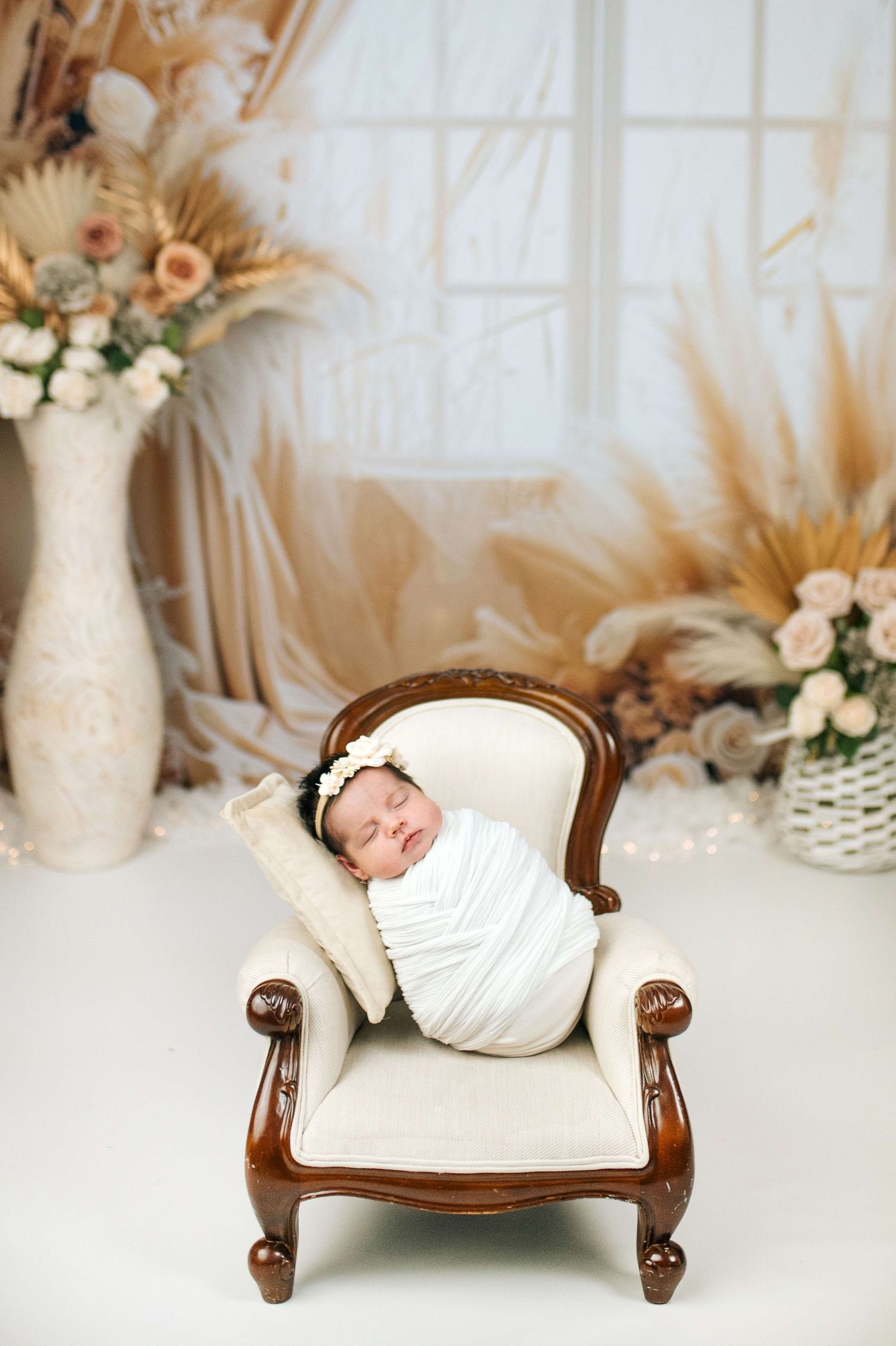Sleeping newborn baby swaddled in white wrap resting on a miniature cream armchair with floral decor backdrop.