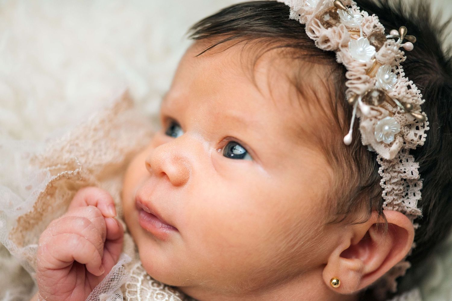 Newborn baby girl with dark hair wearing a floral lace headband and gold earring, looking upward.