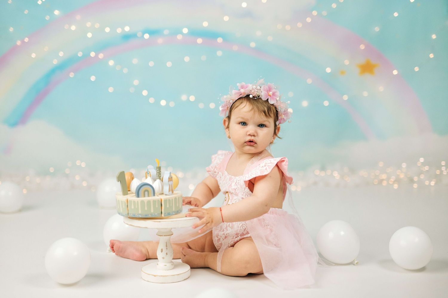 Baby girl in pink romper sits beside a rainbow cake during a first birthday smash cake photo session.