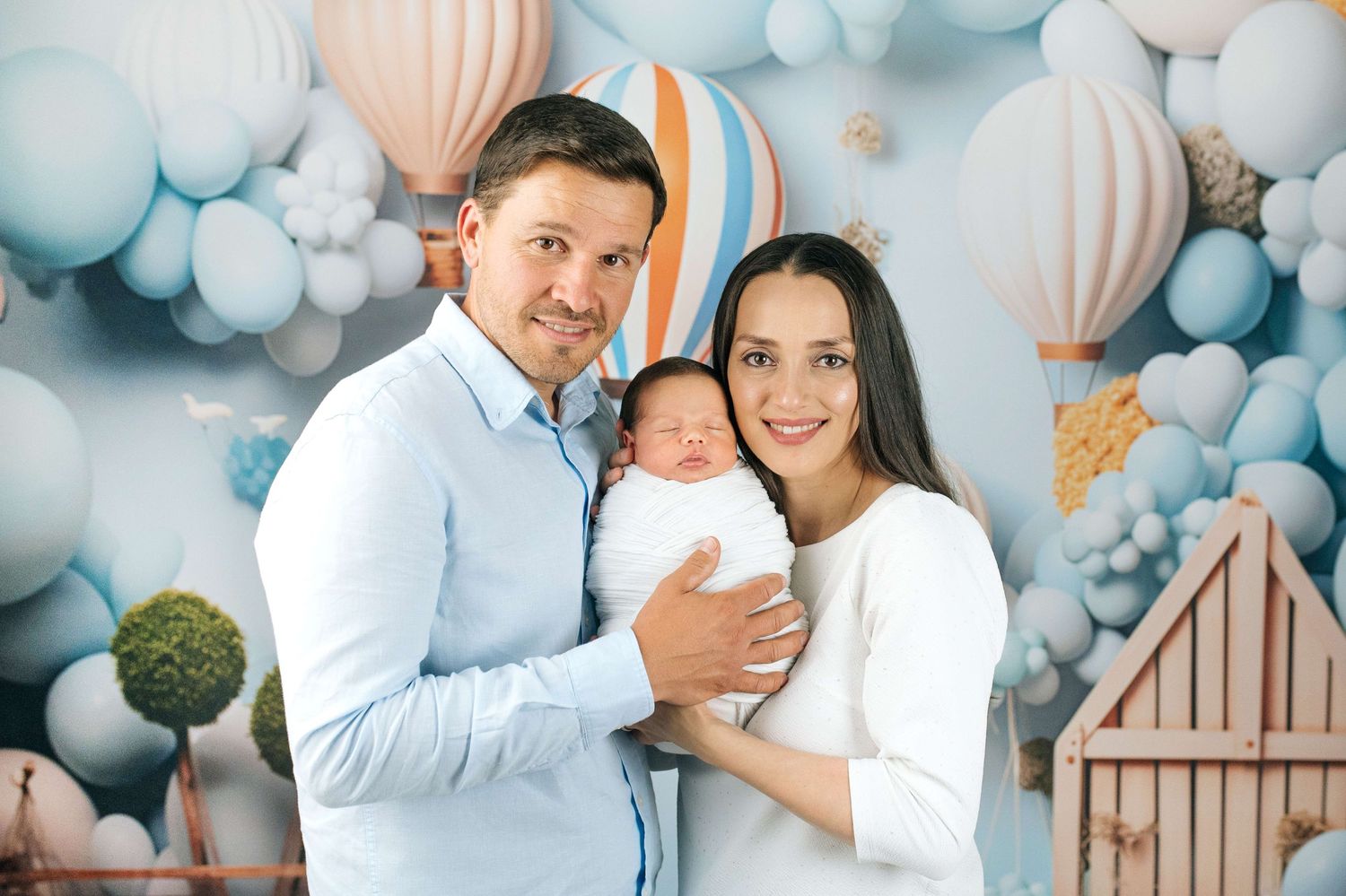 Happy couple holding newborn baby in front of blue balloon and hot air balloon themed backdrop.