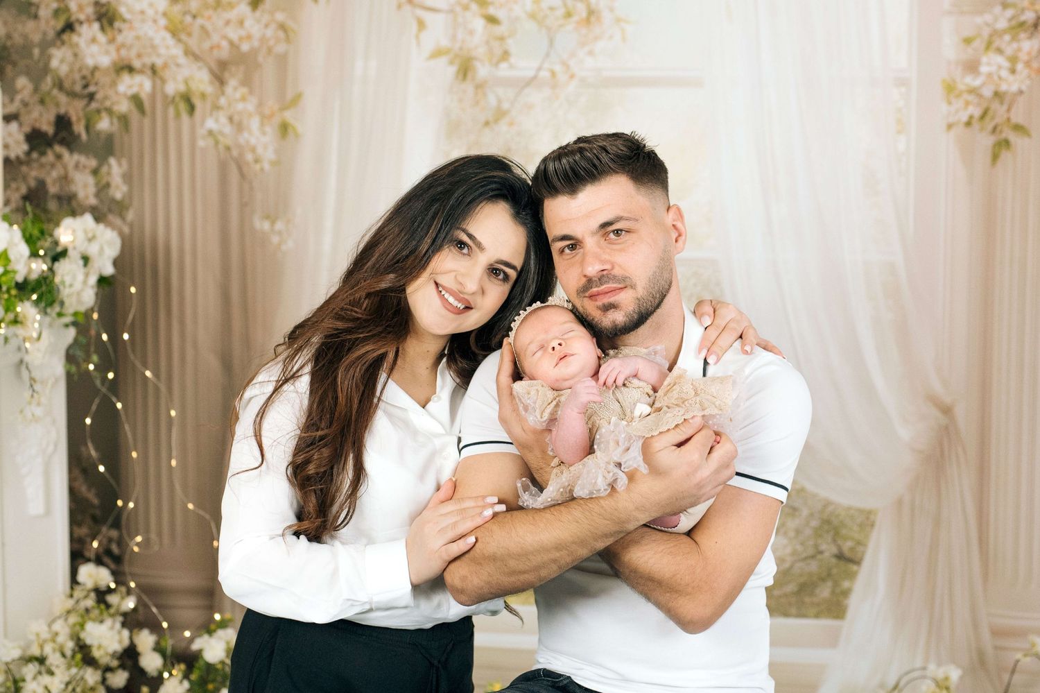 Smiling couple in white tops holding their newborn baby in a softly lit floral photography studio.