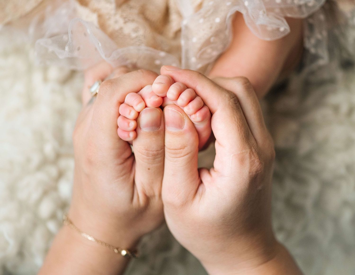 Adult hands gently cradling tiny newborn baby feet, with infant in lace dress on soft white background.