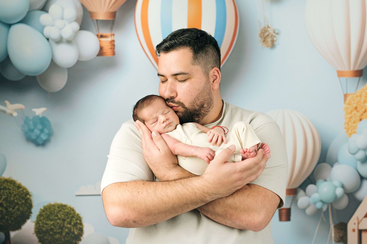 Father tenderly kisses sleeping newborn baby in decorated nursery with blue balloons and hot air balloon backdrop.