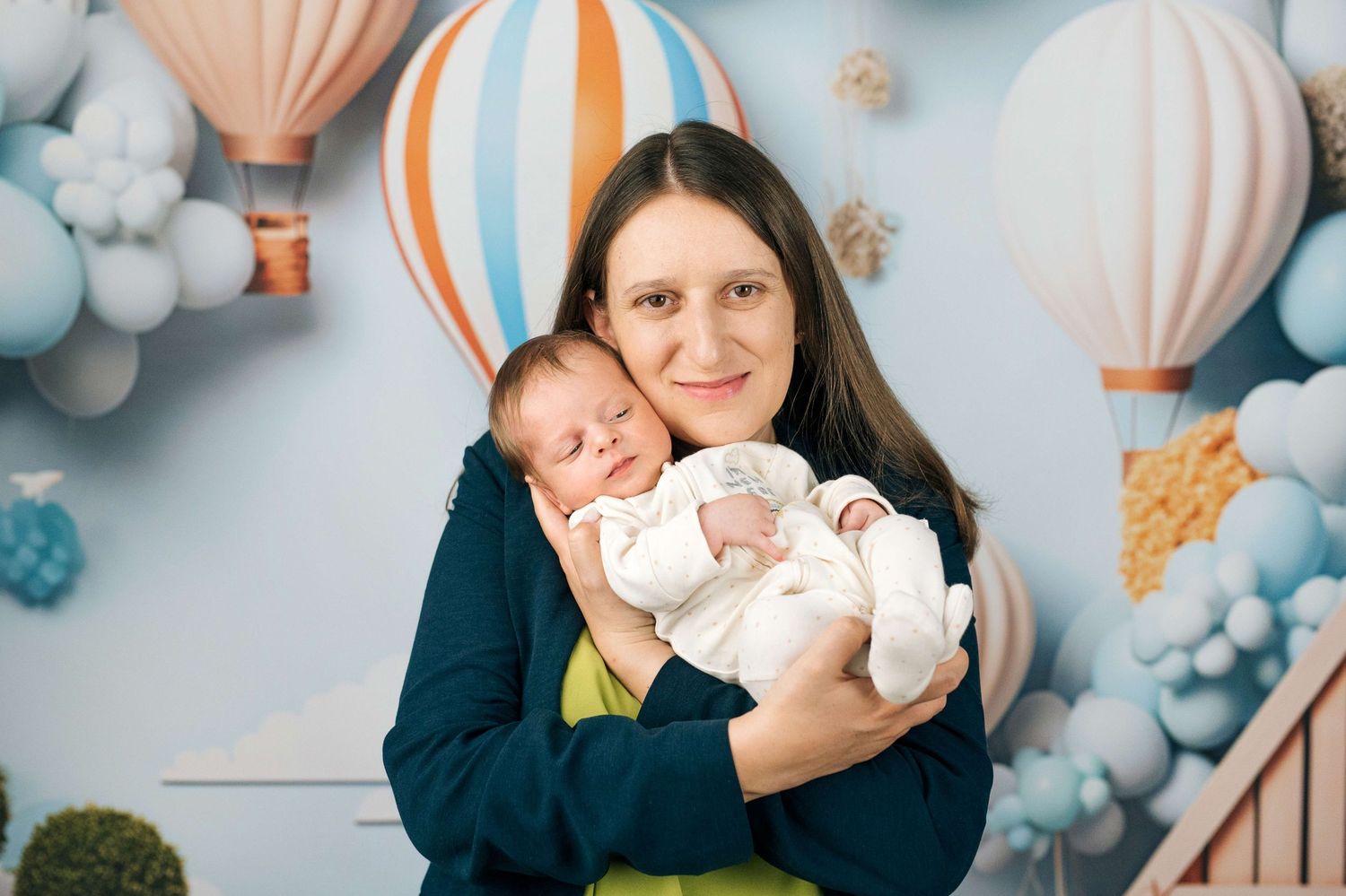 A smiling mother holds two sleeping newborn babies in white outfits against a colorful hot air balloon backdrop.