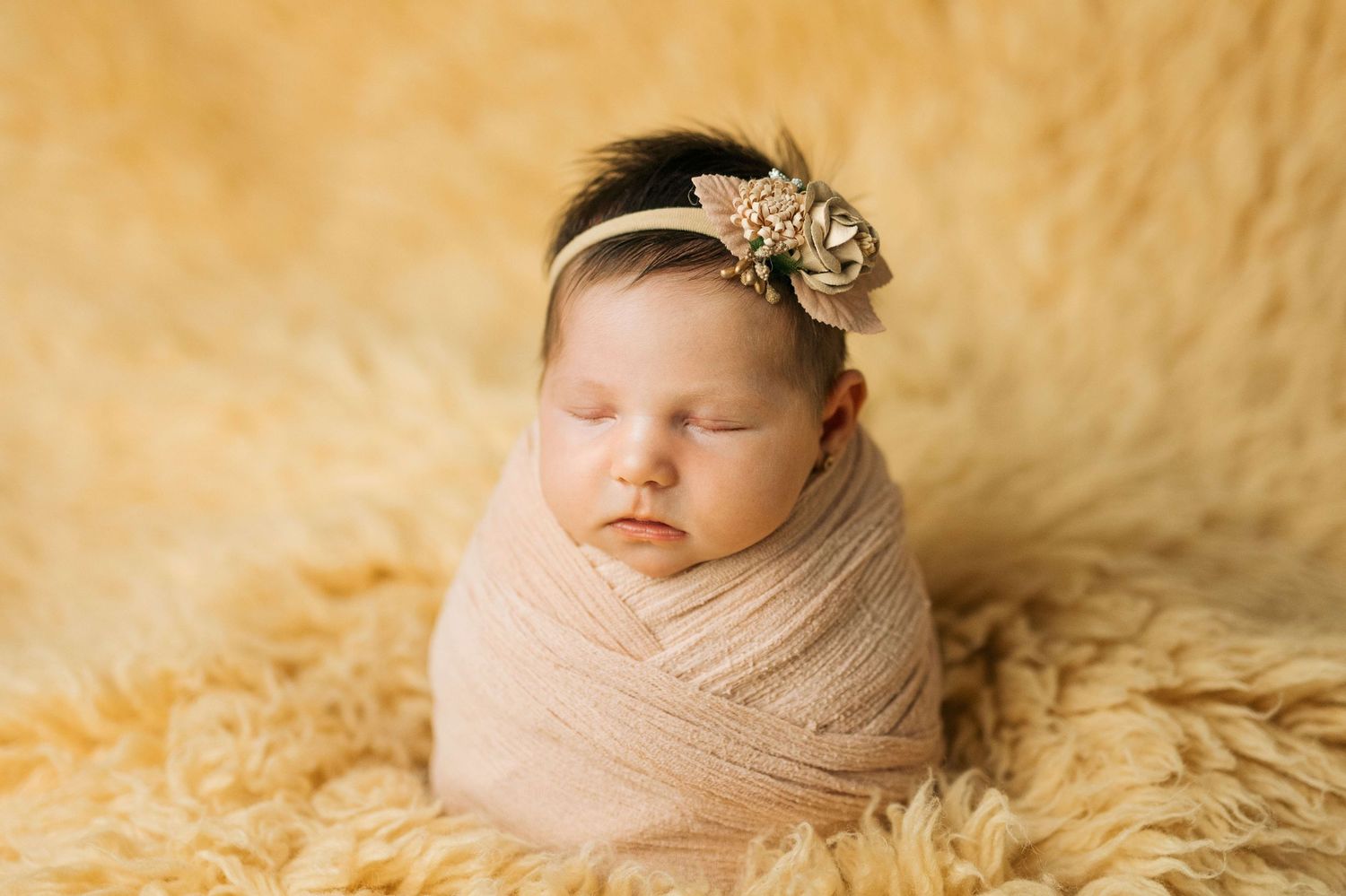 Newborn baby girl swaddled in pink wrap with floral headband sleeping on fluffy yellow fur backdrop.