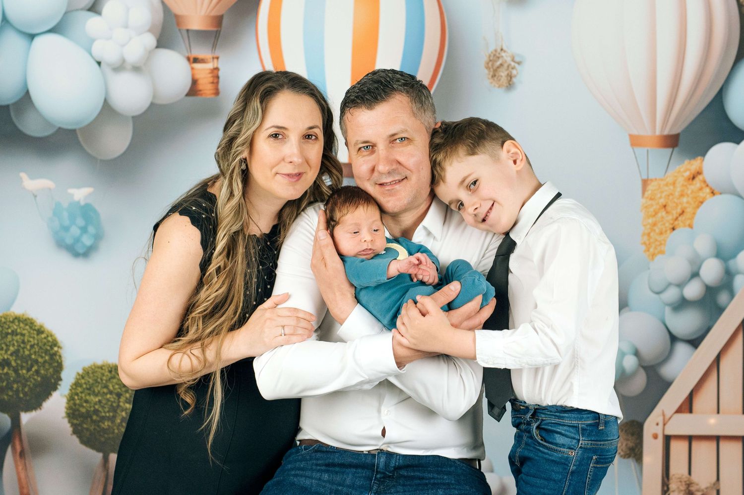 Family of four posing for newborn photo shoot with hot air balloon backdrop, parents holding baby with older son nearby.