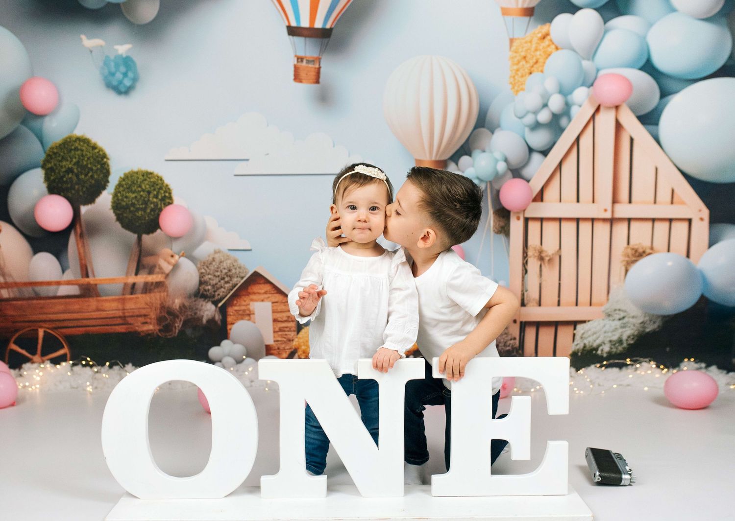 Two young children pose in front of a balloon backdrop with large white ONE letters for a first birthday photo.