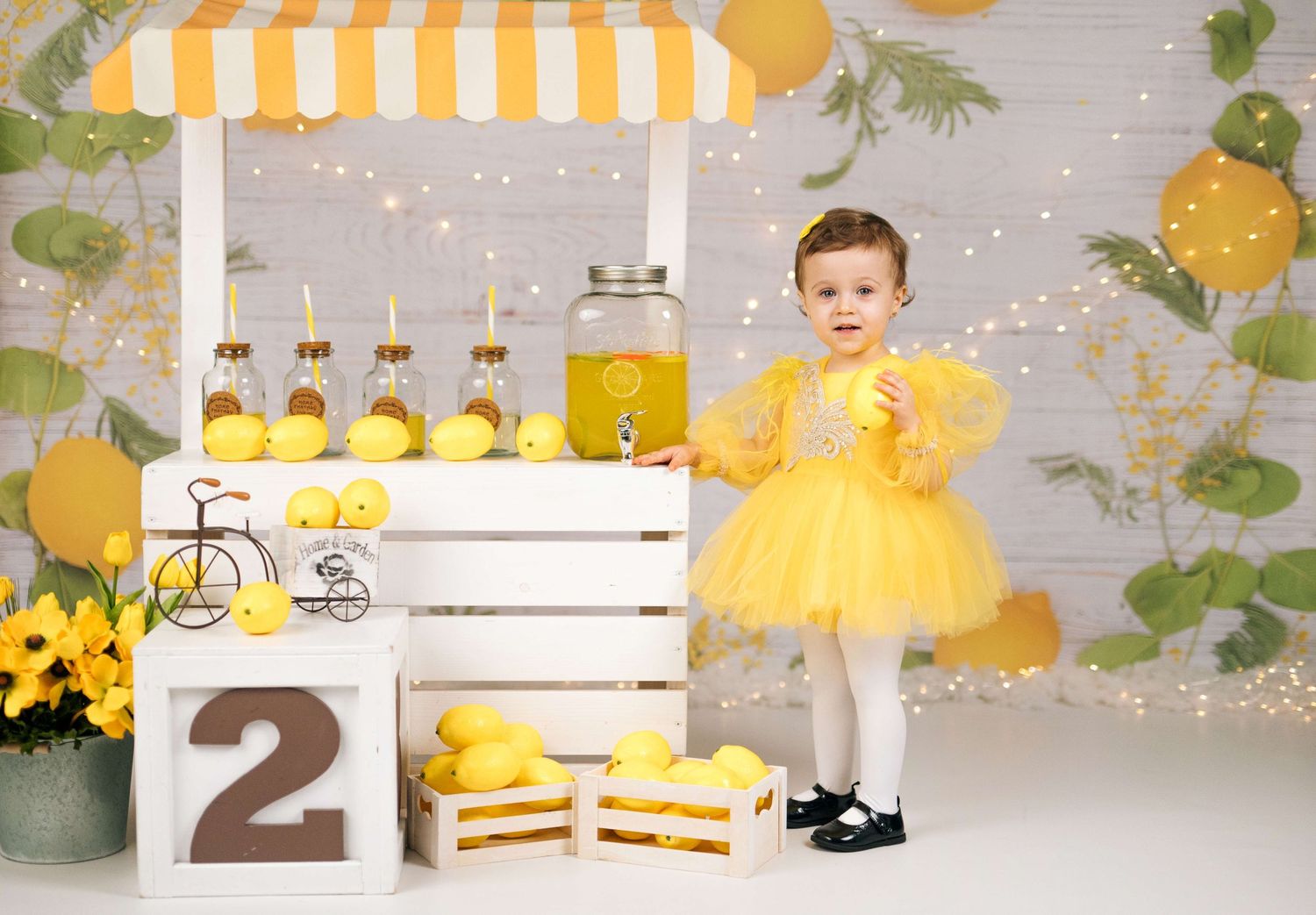 A toddler girl in a yellow tutu dress stands beside a lemonade stand decorated with lemons, next to a number 2 block.