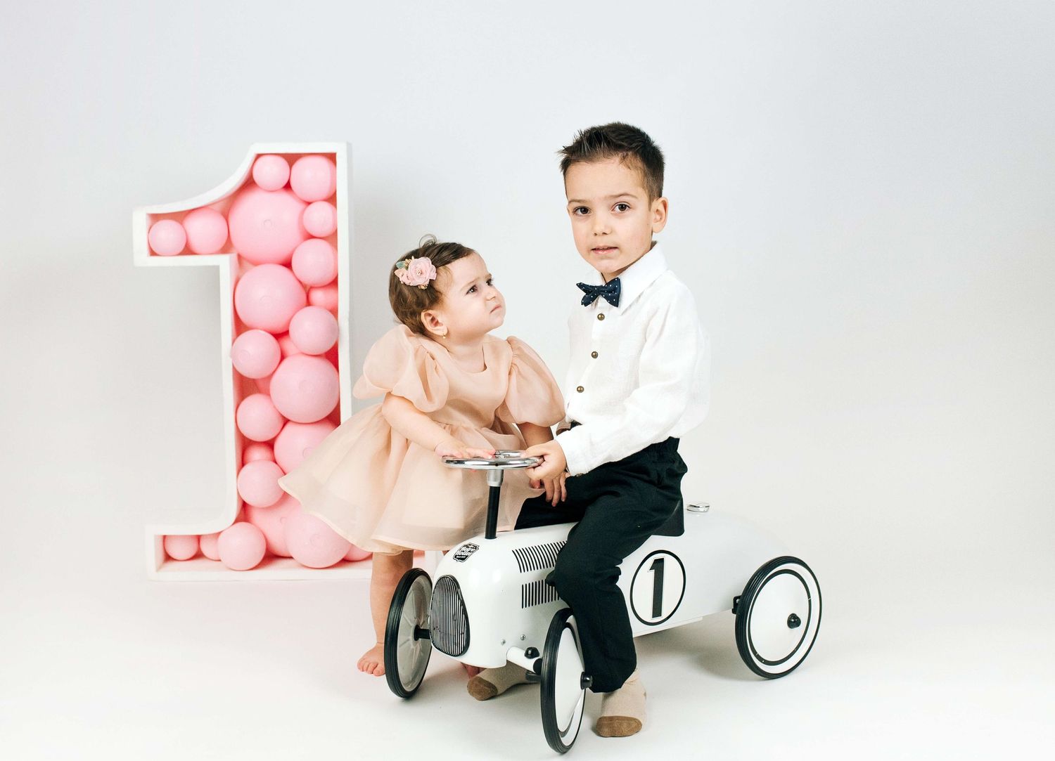 Two young children pose with a large pink balloon number one, a baby girl in pink dress and boy in tuxedo on toy car.