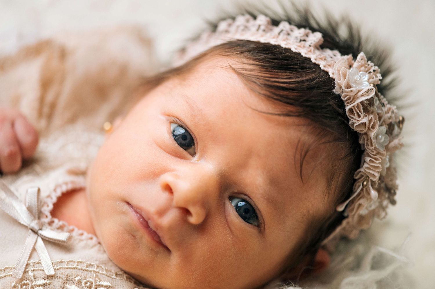 Newborn baby girl with dark hair wearing a delicate lace headband and outfit, lying on a soft white blanket.