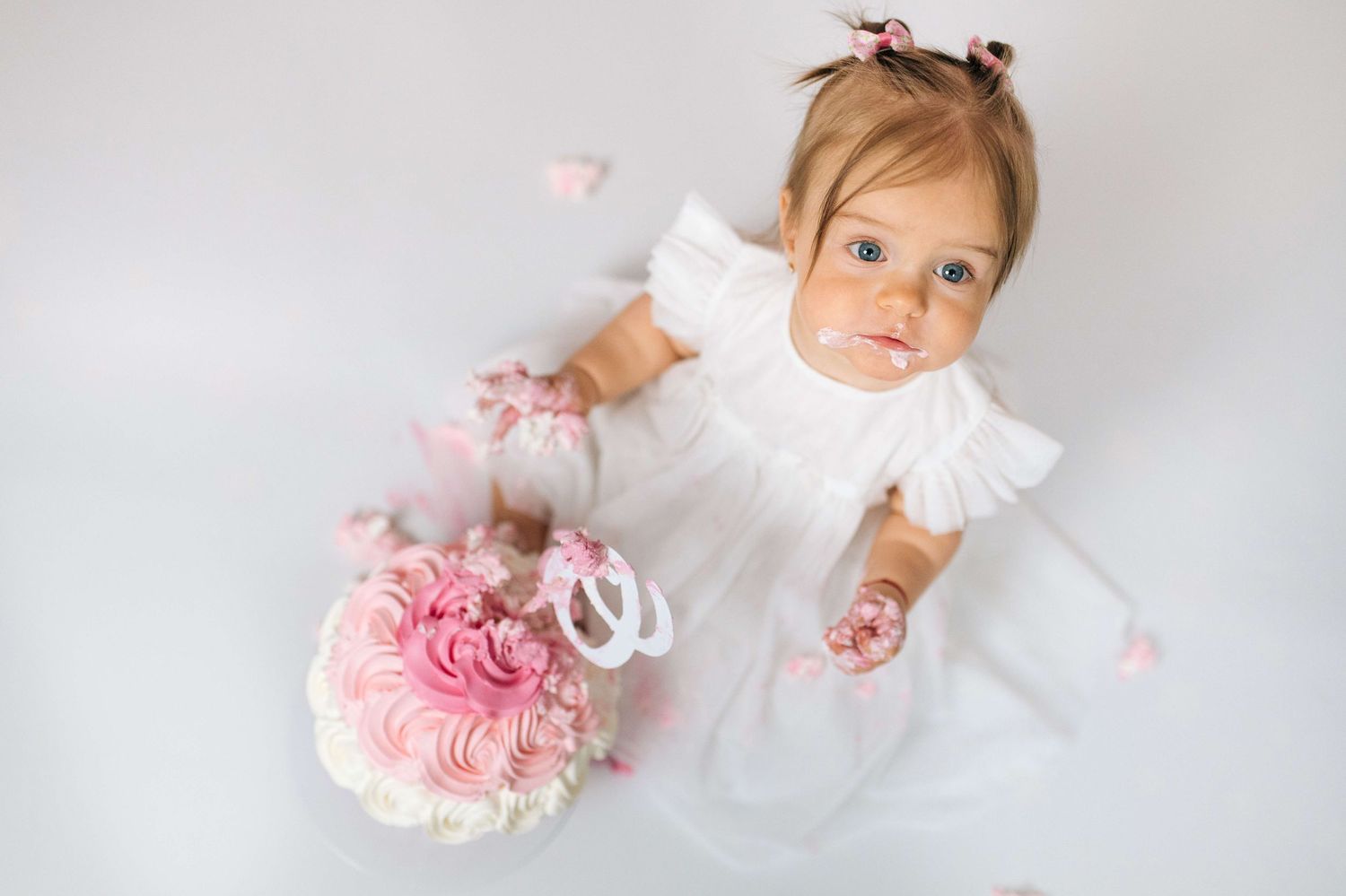 Baby girl in white dress sits on floor with pink floral cake, celebrating her first birthday.