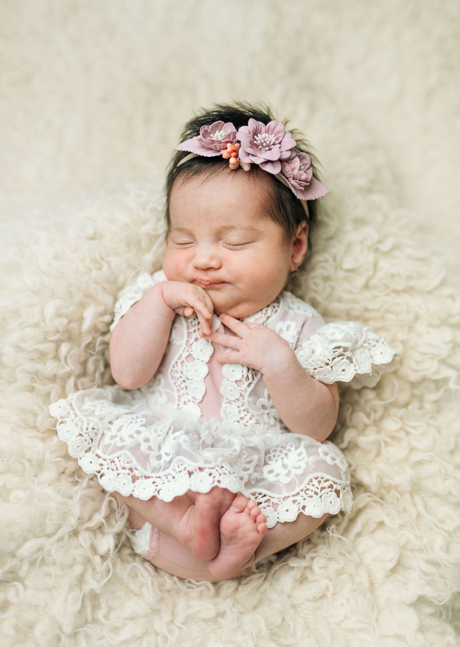 Sleeping newborn baby girl in white lace dress and pink floral headband on fluffy cream backdrop.