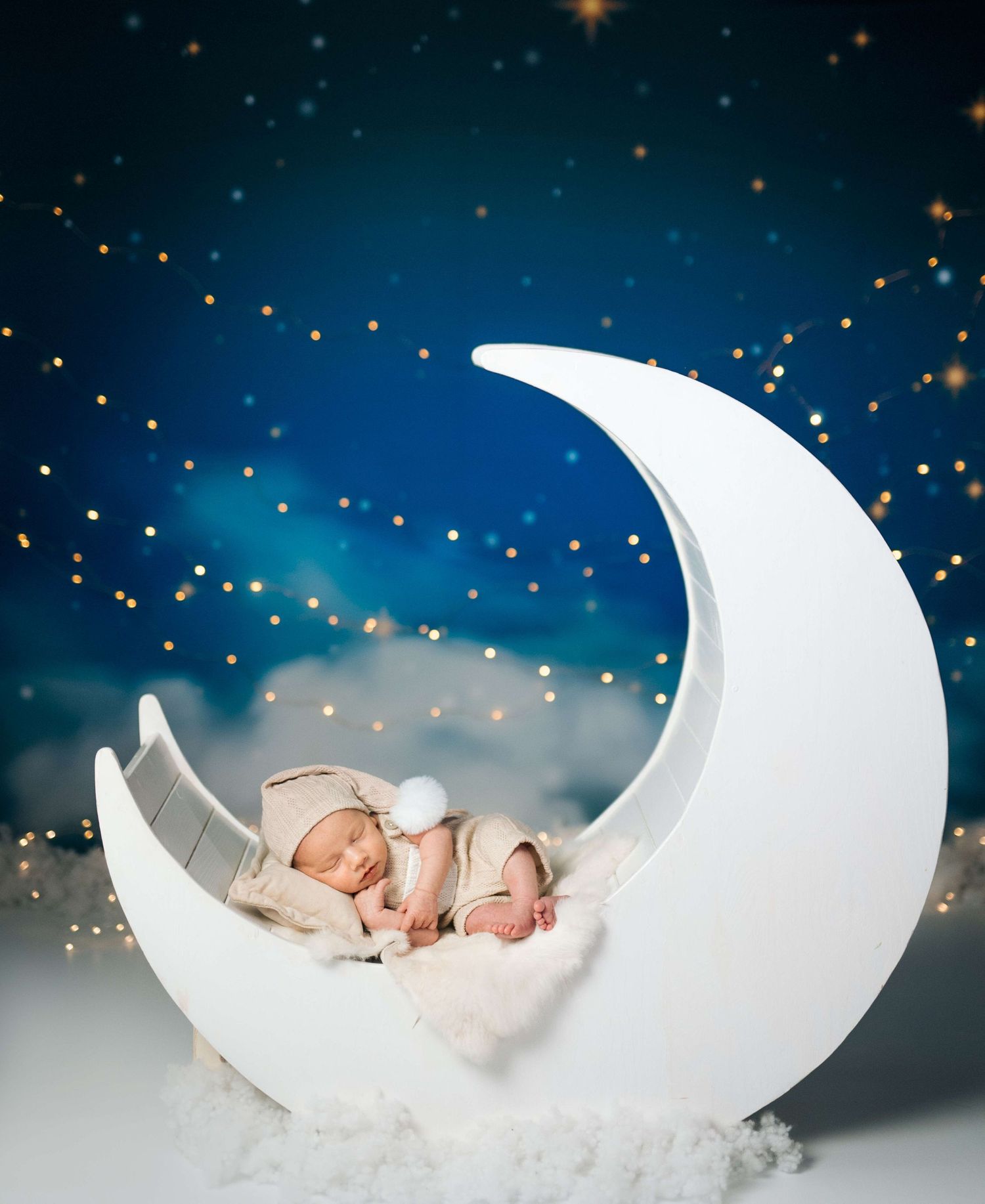 Newborn baby sleeping peacefully on a white crescent moon prop against a dreamy starry night sky backdrop.