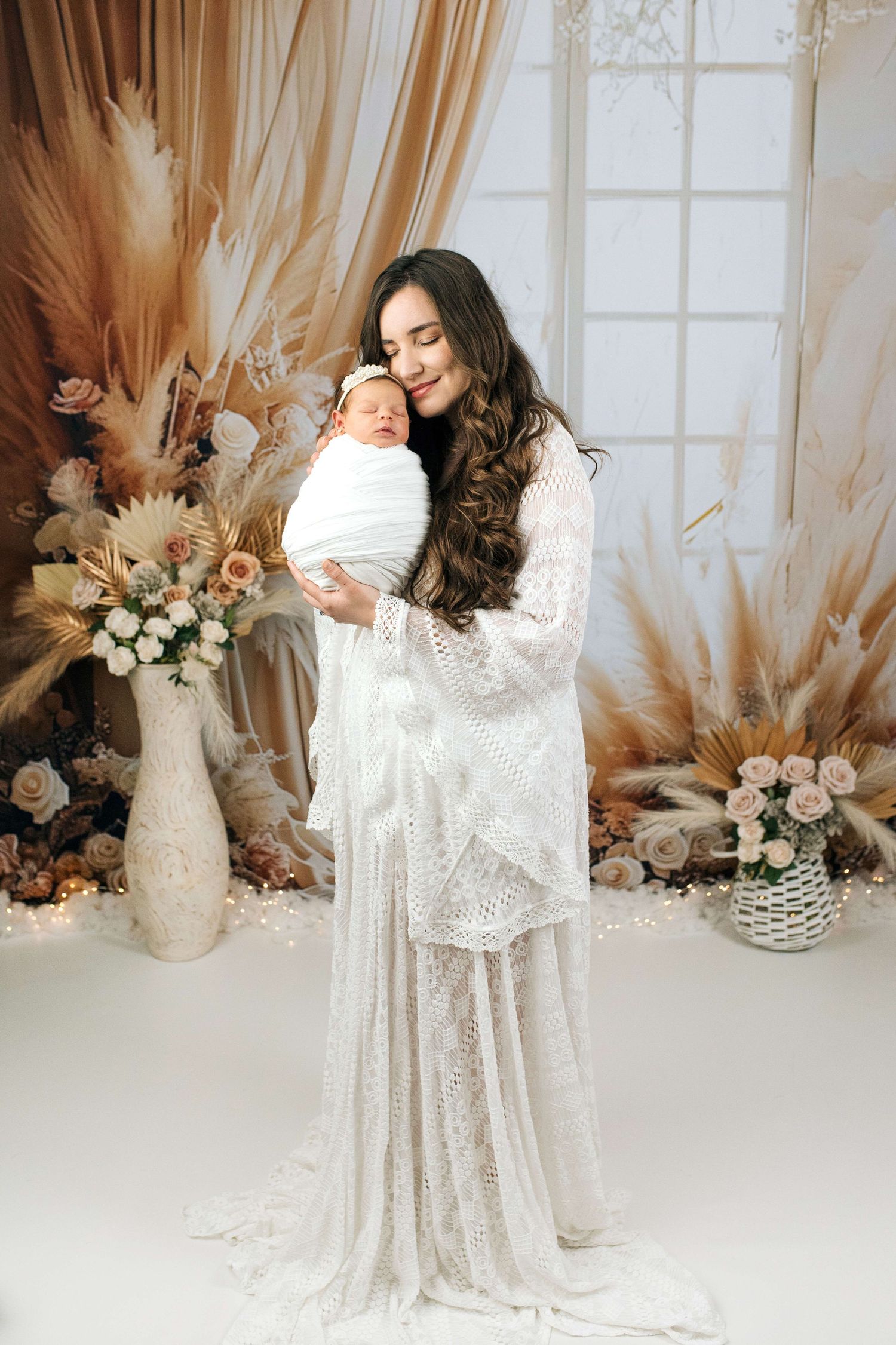 A mother in a white lace gown tenderly holds her swaddled newborn in a boho-styled studio with dried florals.
