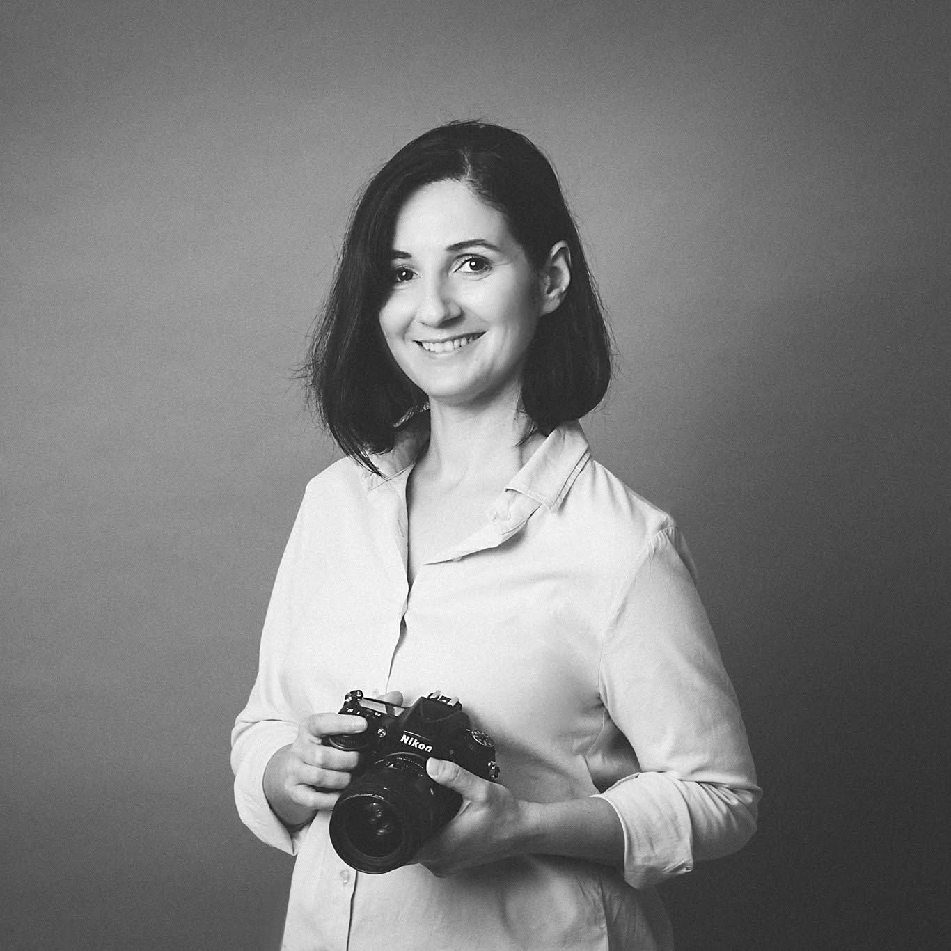 Black and white portrait of a smiling woman holding a Nikon camera, wearing a white blouse.