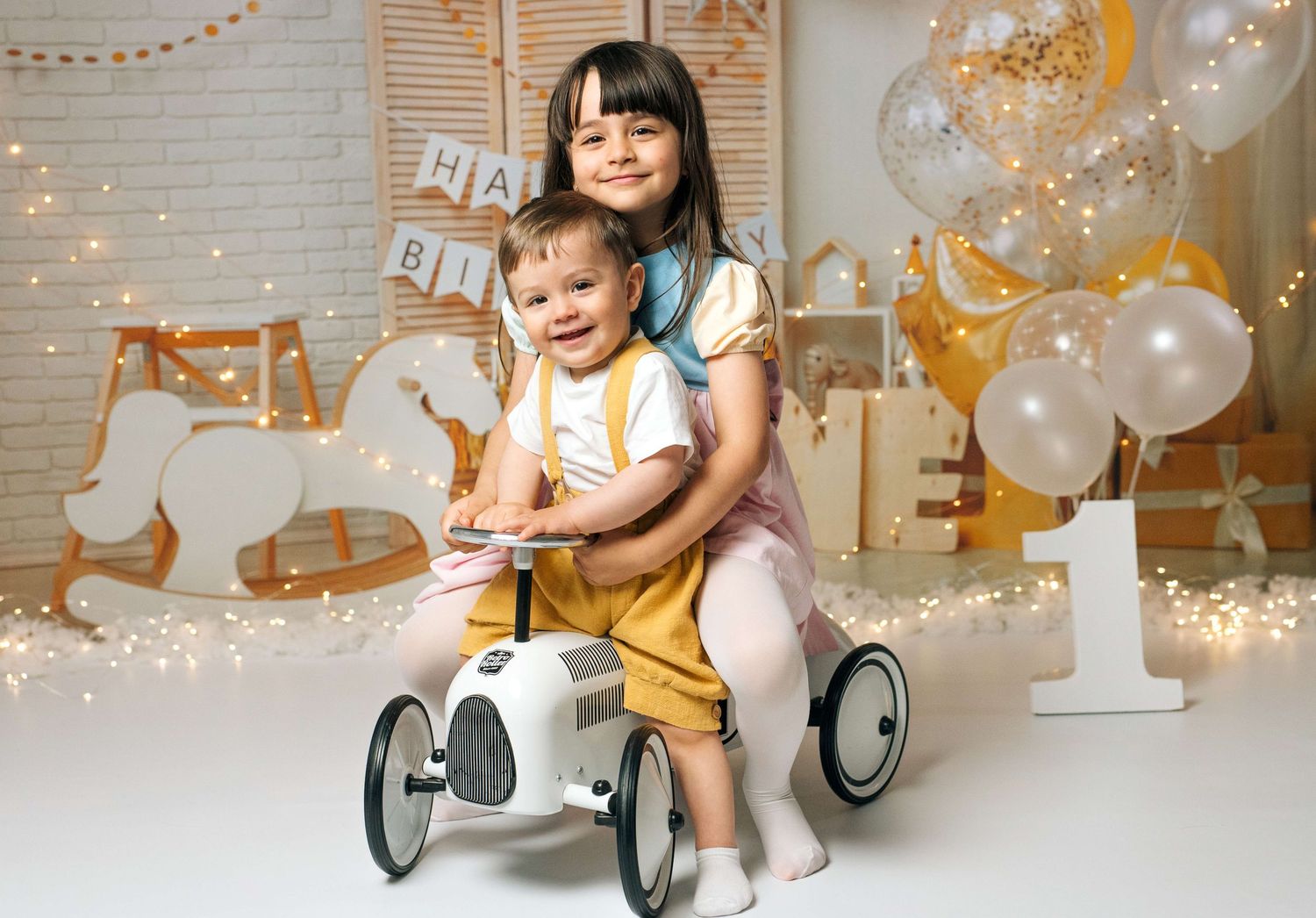 Two children pose on a vintage toy car with balloons and fairy lights in a festive birthday photo studio setting.