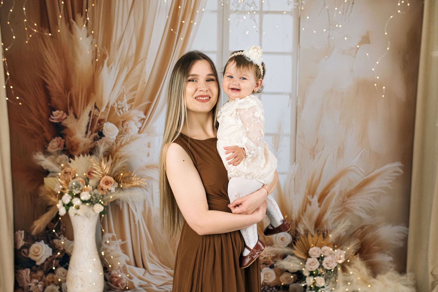 A mother in a brown dress holds her baby girl in a white dress against a boho floral backdrop.