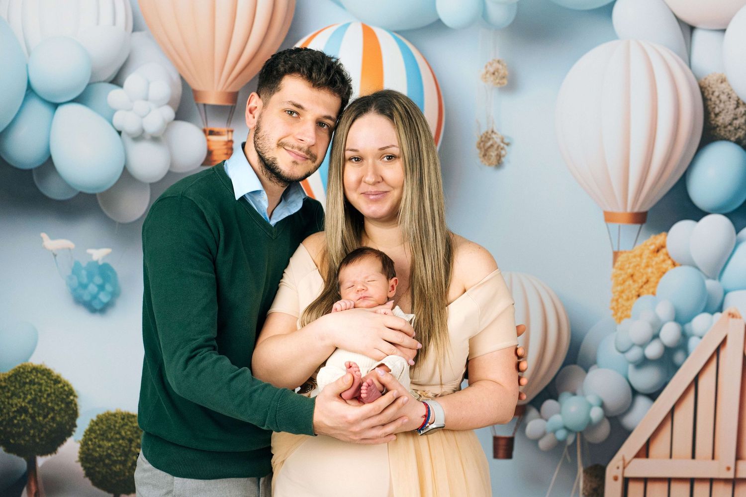 Young couple smiling and holding their newborn baby in front of a blue balloon and hot air balloon backdrop.