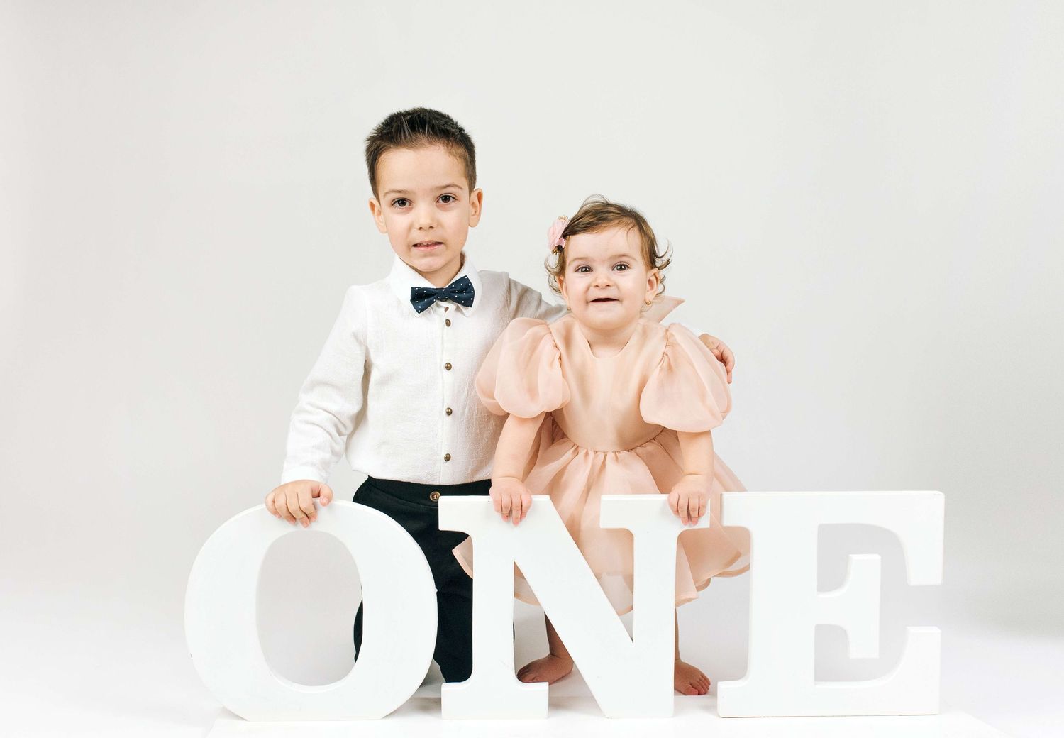 Two dressed-up children pose with large white ONE letters for a first birthday photo session.