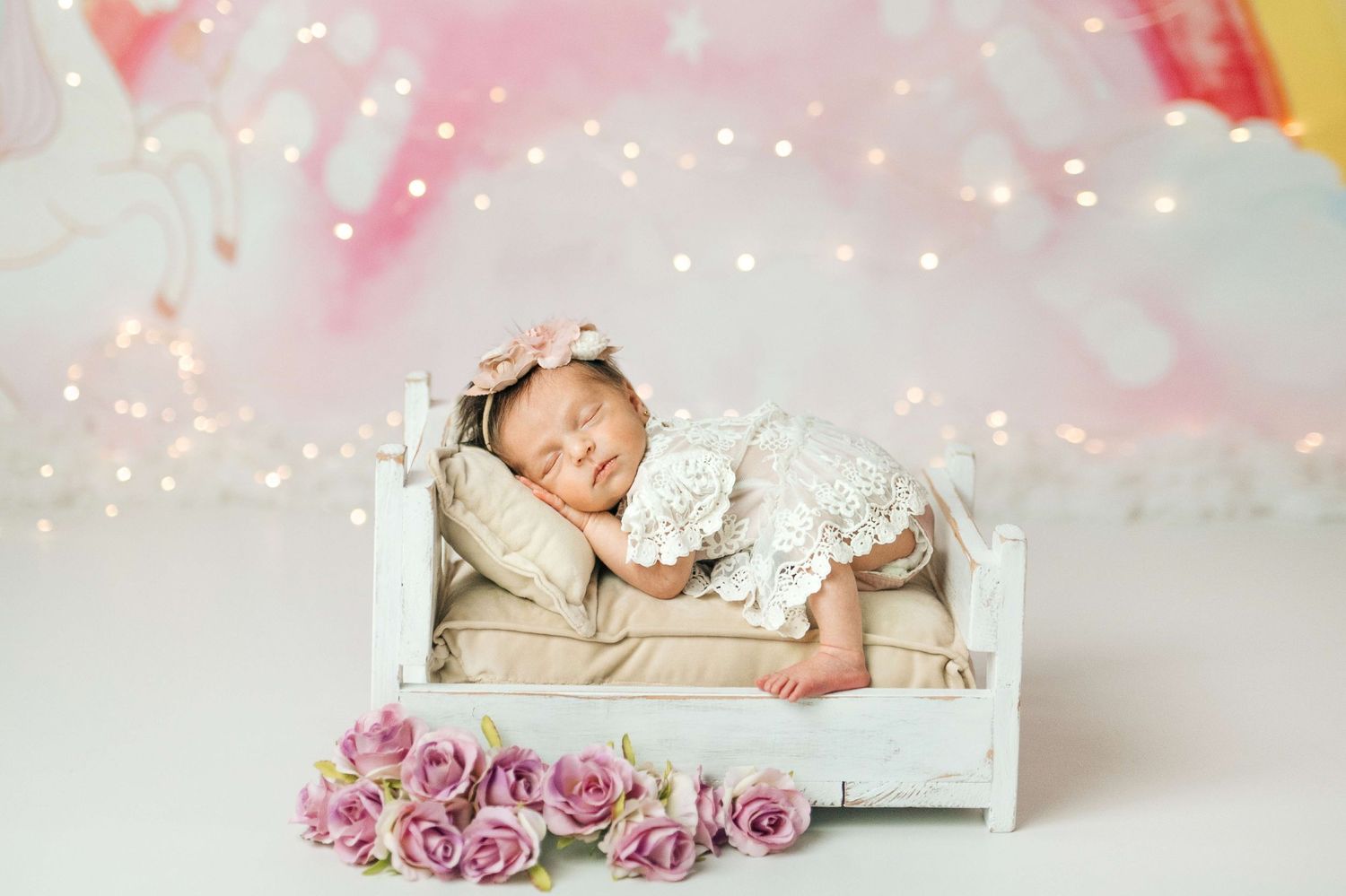 Newborn baby girl sleeping peacefully on a tiny white bed with pink roses and bokeh lights in background.