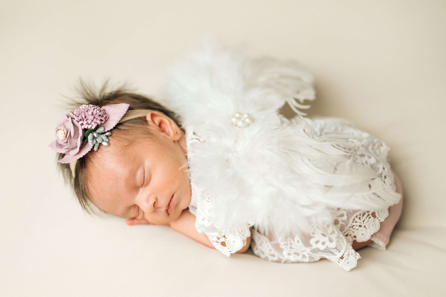 Sleeping newborn baby girl in white lace outfit and floral headband on cream background.