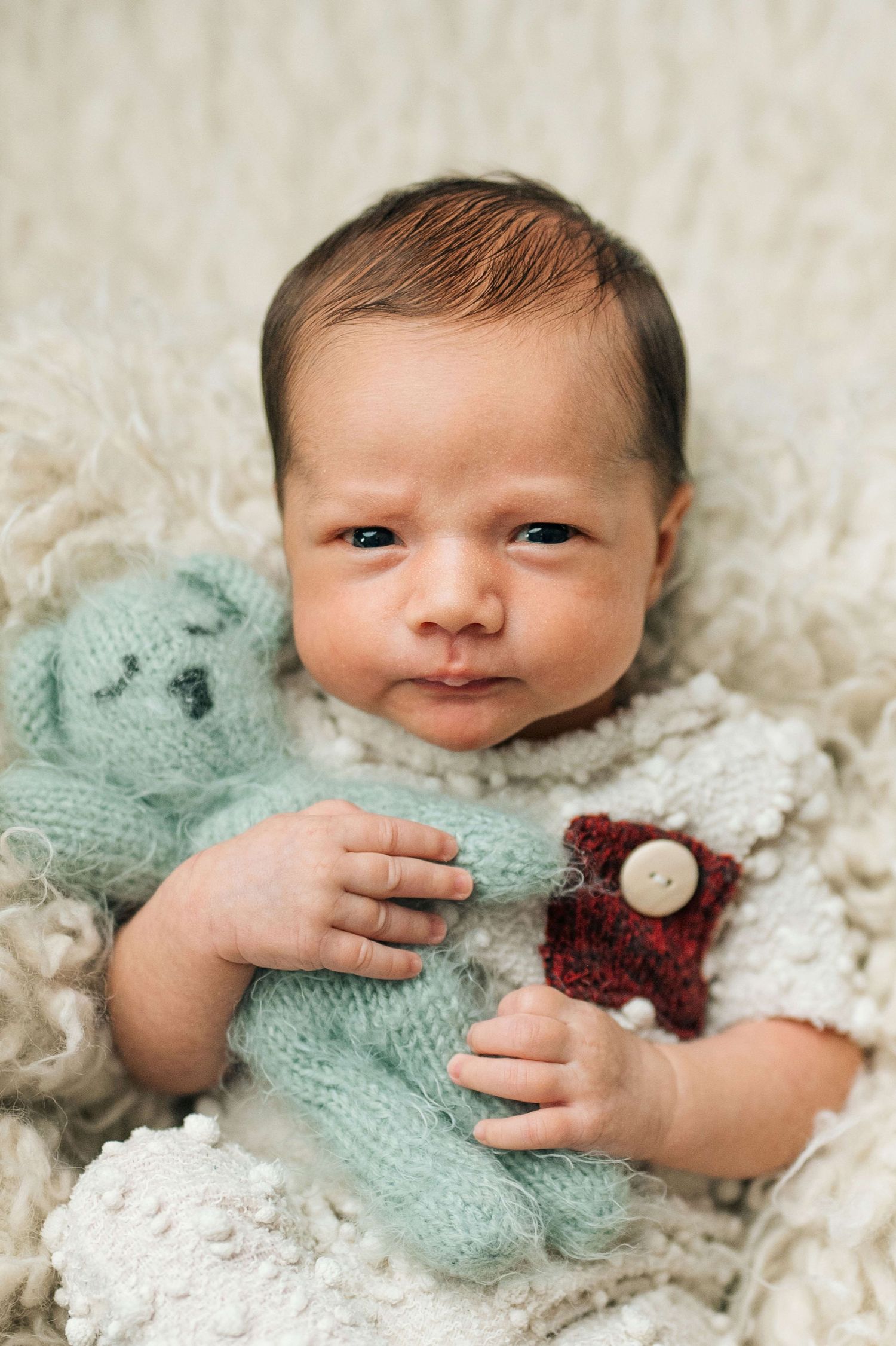 Newborn baby lying on fluffy white blanket holding two small knitted teddy bears, one mint green and one burgundy red.