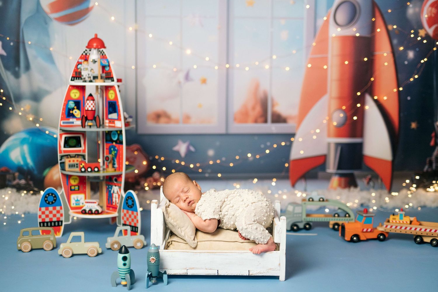 Sleeping newborn baby on white mini bed surrounded by toy rockets, cars, and twinkling lights in a space-themed nursery.